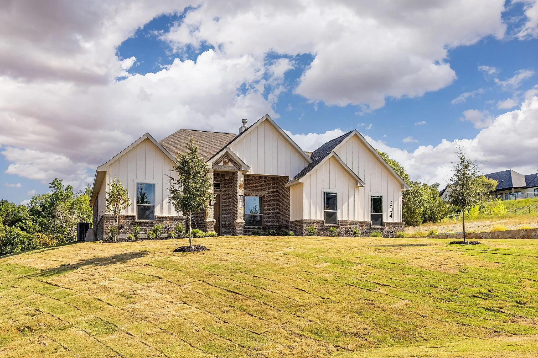 Board and batten siding, brick facade, stone accents, gabled rooflines, and a sloped lawn