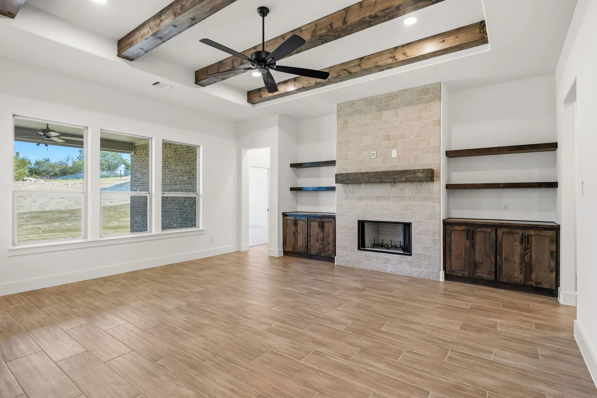 Tray ceiling with exposed wood beams, tile-surround fireplace with wood mantle, built-in shelving, wood-finish flooring, and multiple windows providing natural illumination