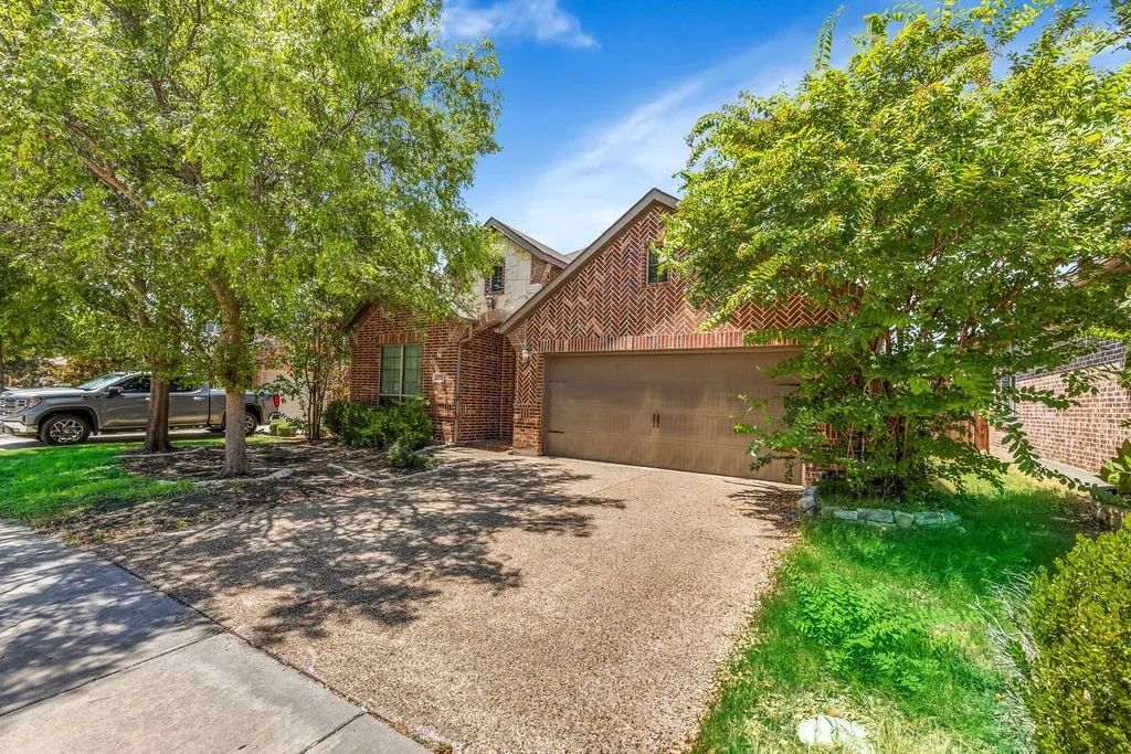 Brick exterior featuring a two-car garage, mature trees, and a textured driveway