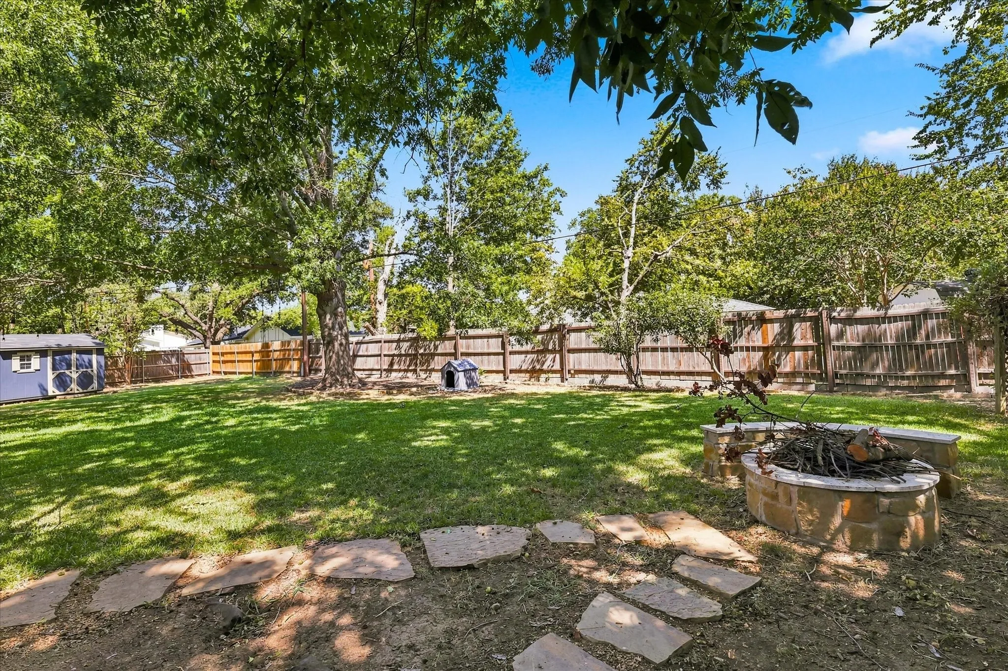 Expansive backyard featuring a stone fire pit, natural stone stepping stones, a wooden privacy fence, mature trees, and a storage shed