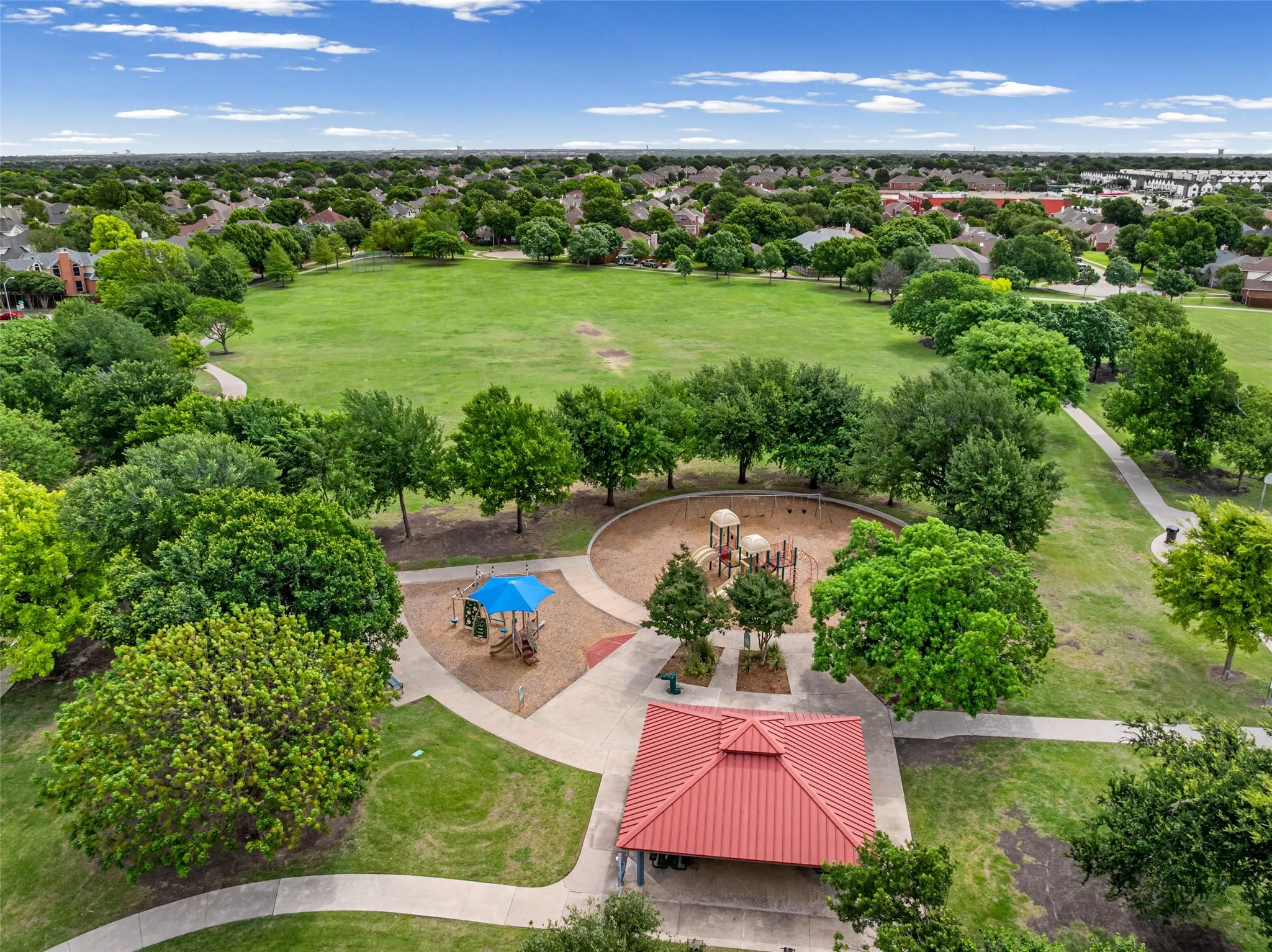 Expansive green space featuring mature trees, a dedicated playground area, paved walking paths, and a covered pavilion