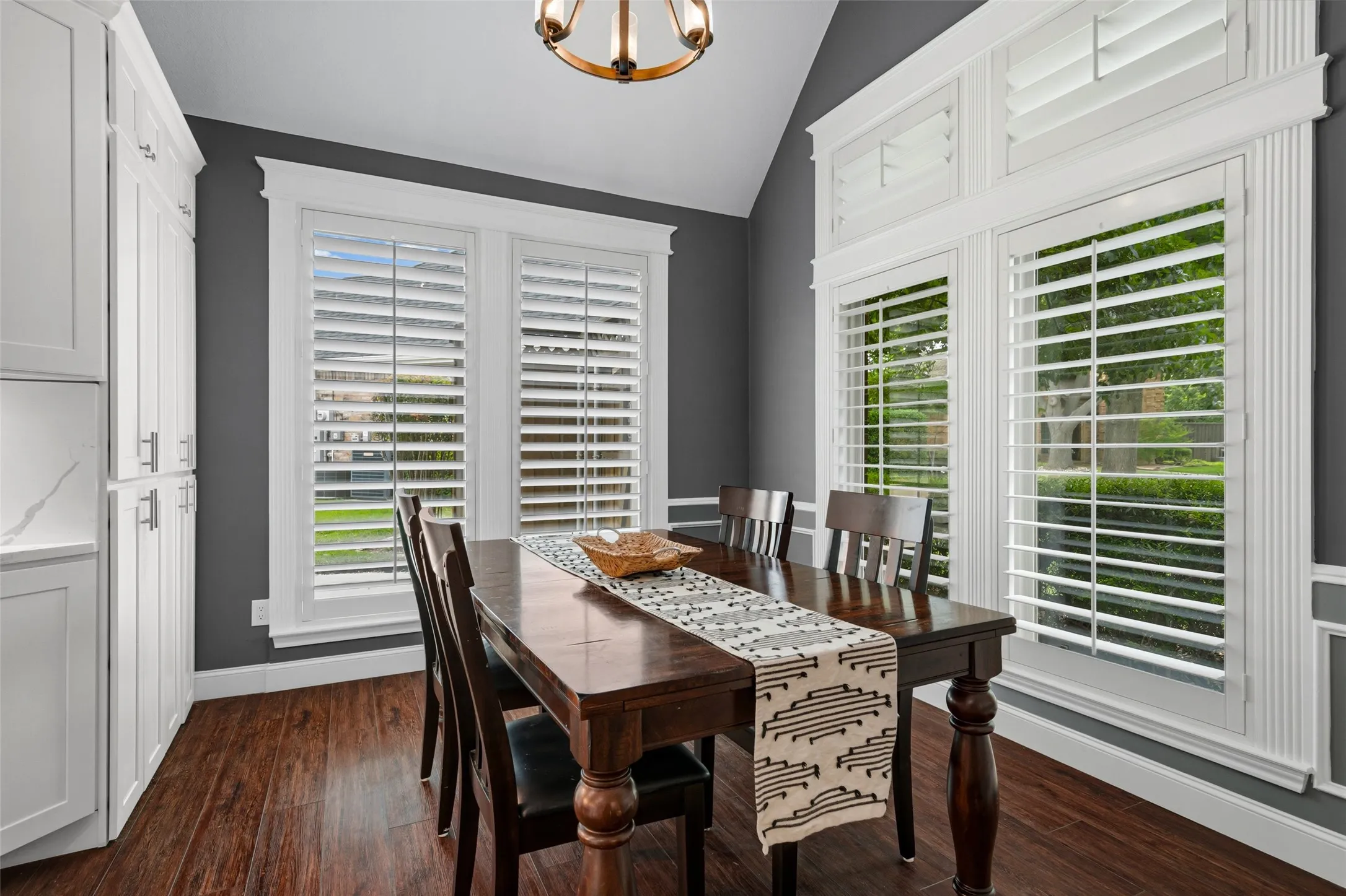 Vaulted ceiling, white cabinetry with silver-tone hardware, wood-finish flooring, white plantation shutters, and decorative wall molding