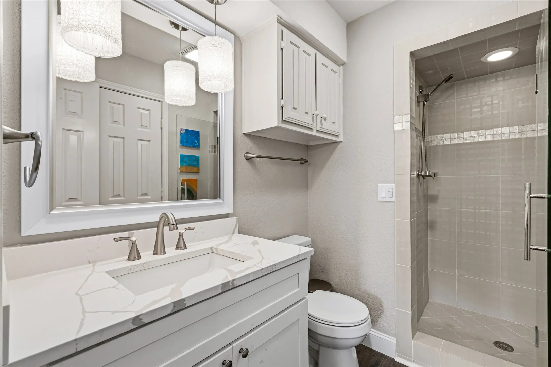 White vanity with a rectangular undermount sink and a light-toned countertop, framed mirror with pendant lighting, and a walk-in shower with tiled walls and a glass door