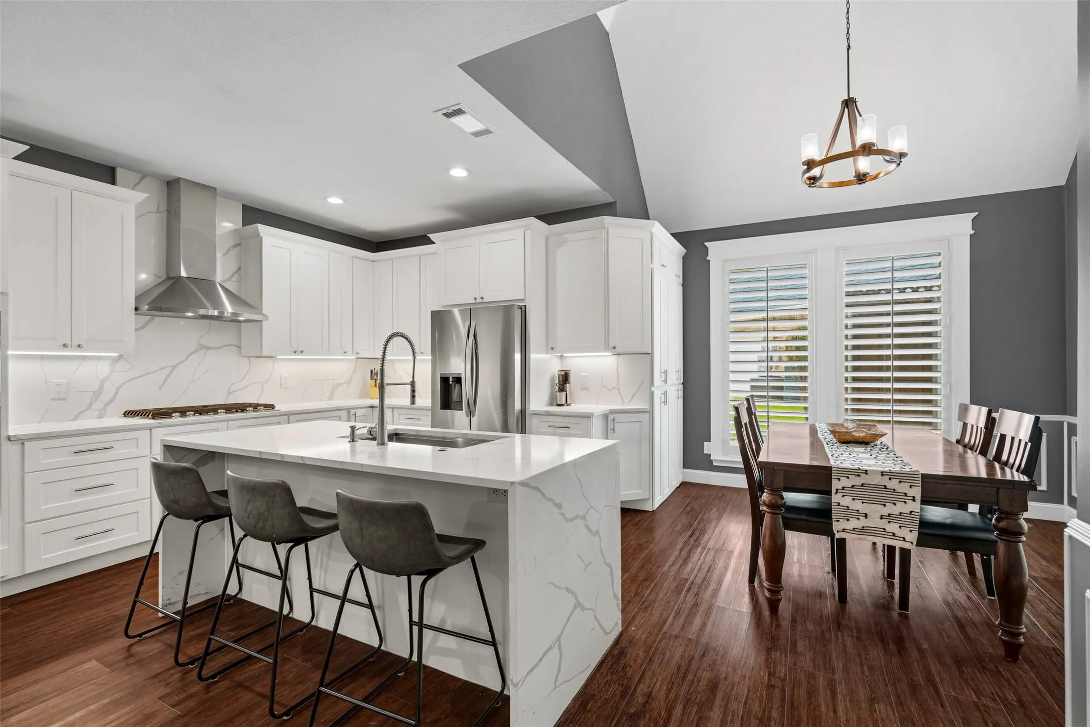 Modern kitchen and dining area featuring white cabinetry, marble-finish island with waterfall edge and seating for three, stainless steel appliances, pendant lighting, wood-finish flooring, and plantation shutters overlooking the dining space with coffered ceiling detail
