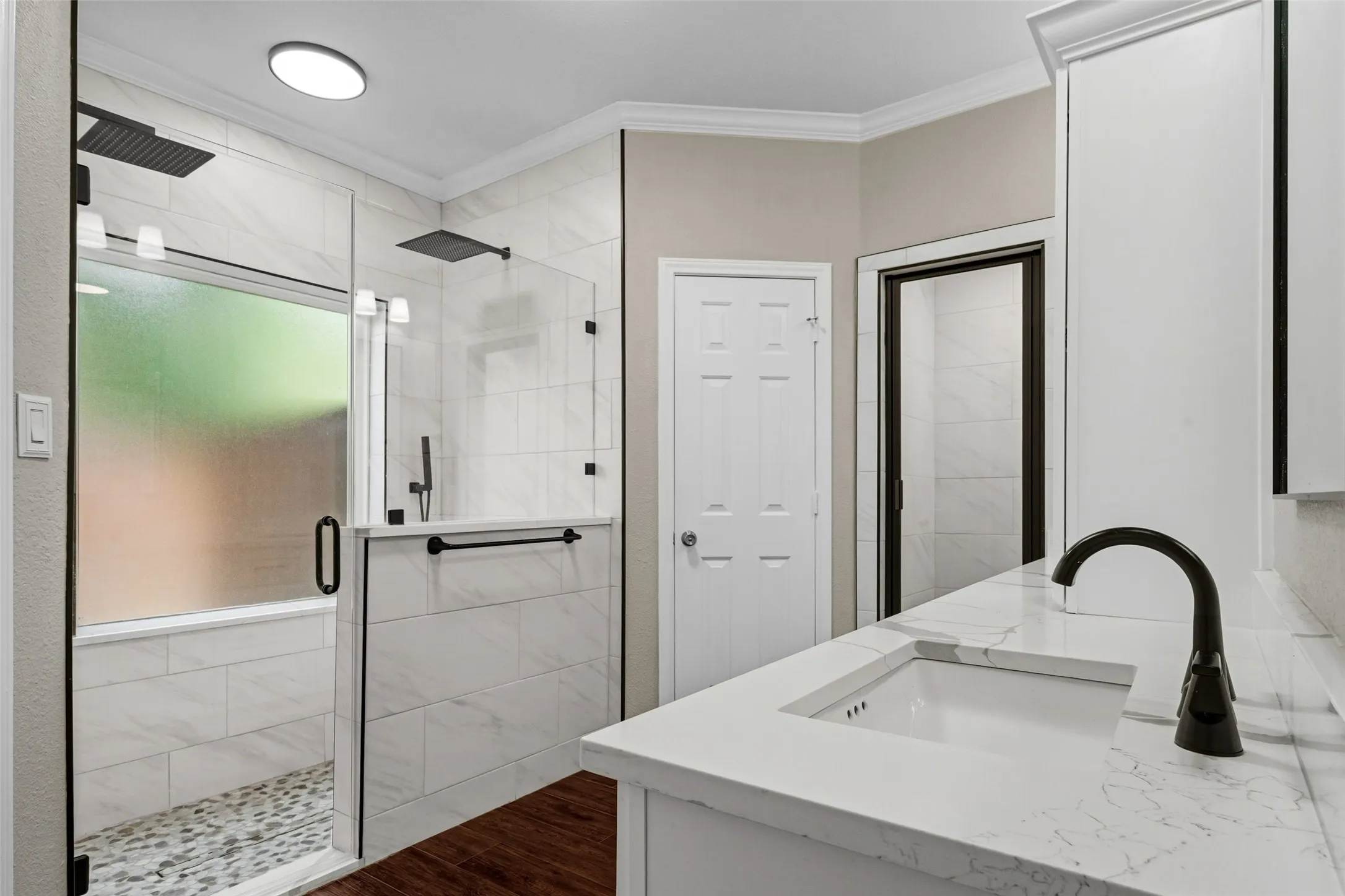 Dual shower heads, glass-enclosed shower with tiled walls and pebble-finish floor, white vanity with integrated sink, black-finish faucet, and wood-finish flooring