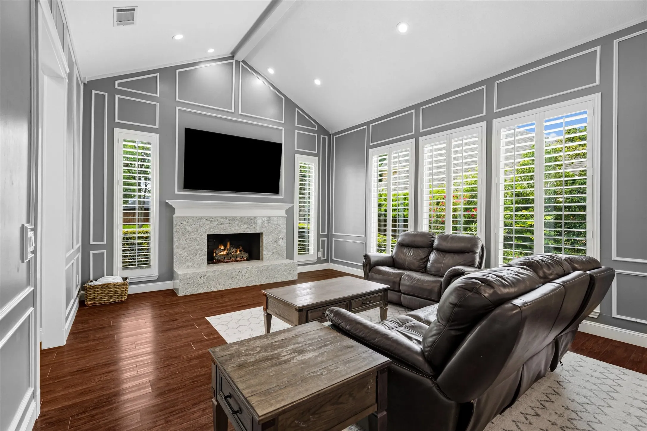 Living area featuring vaulted ceilings, wood-finish flooring, a fireplace with a stone surround, and multiple tall windows with plantation shutters