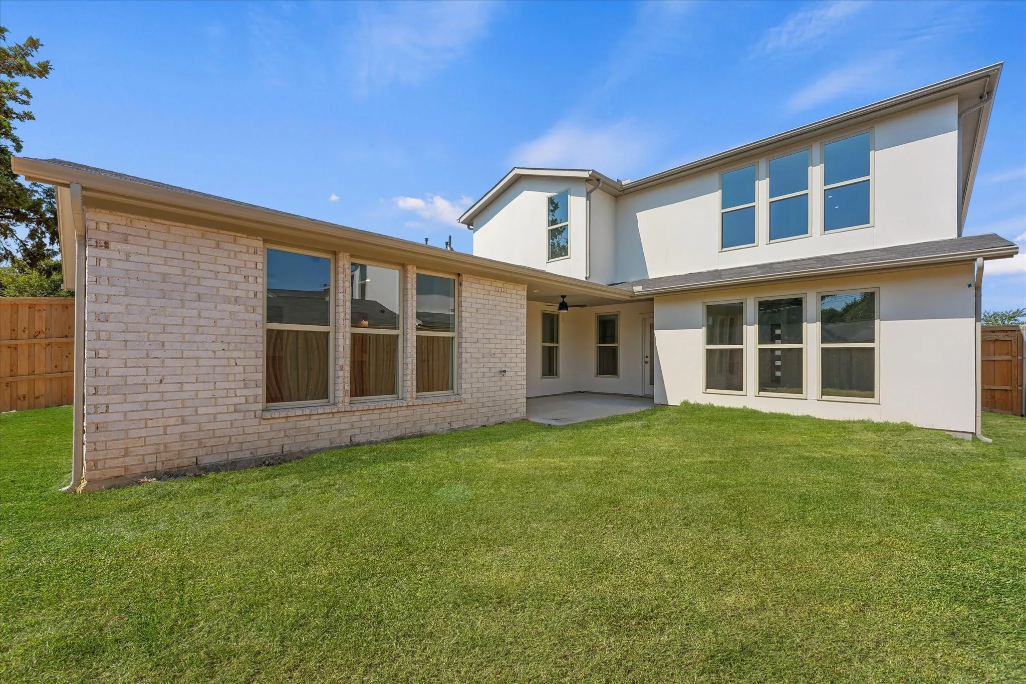 Expansive grass lawn with a covered patio, light-colored brick exterior, stucco siding, and multiple large windows
