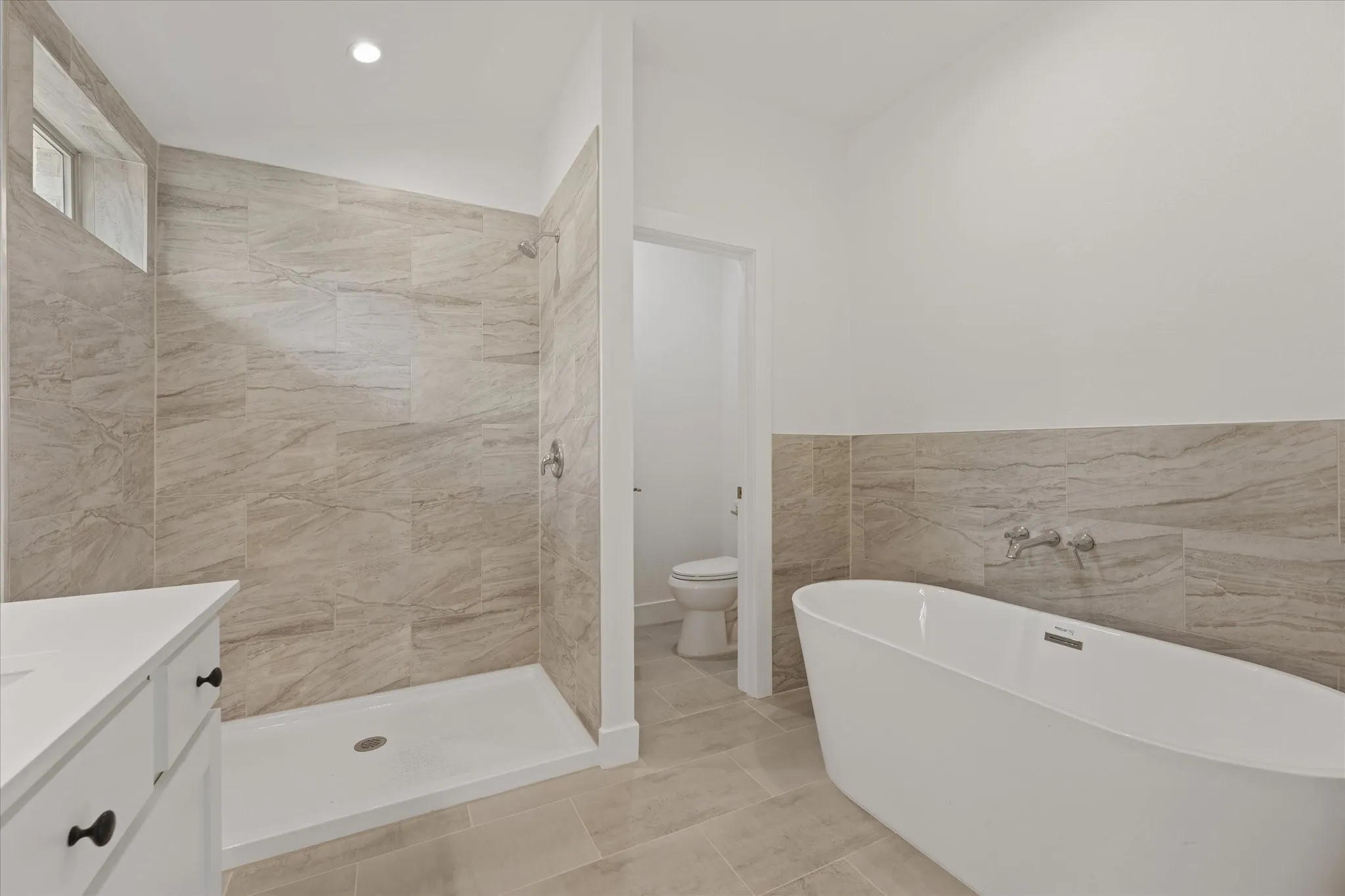 Freestanding soaking tub, tiled shower with transom window, white vanity with dark hardware, and light-toned floor and wall tiling