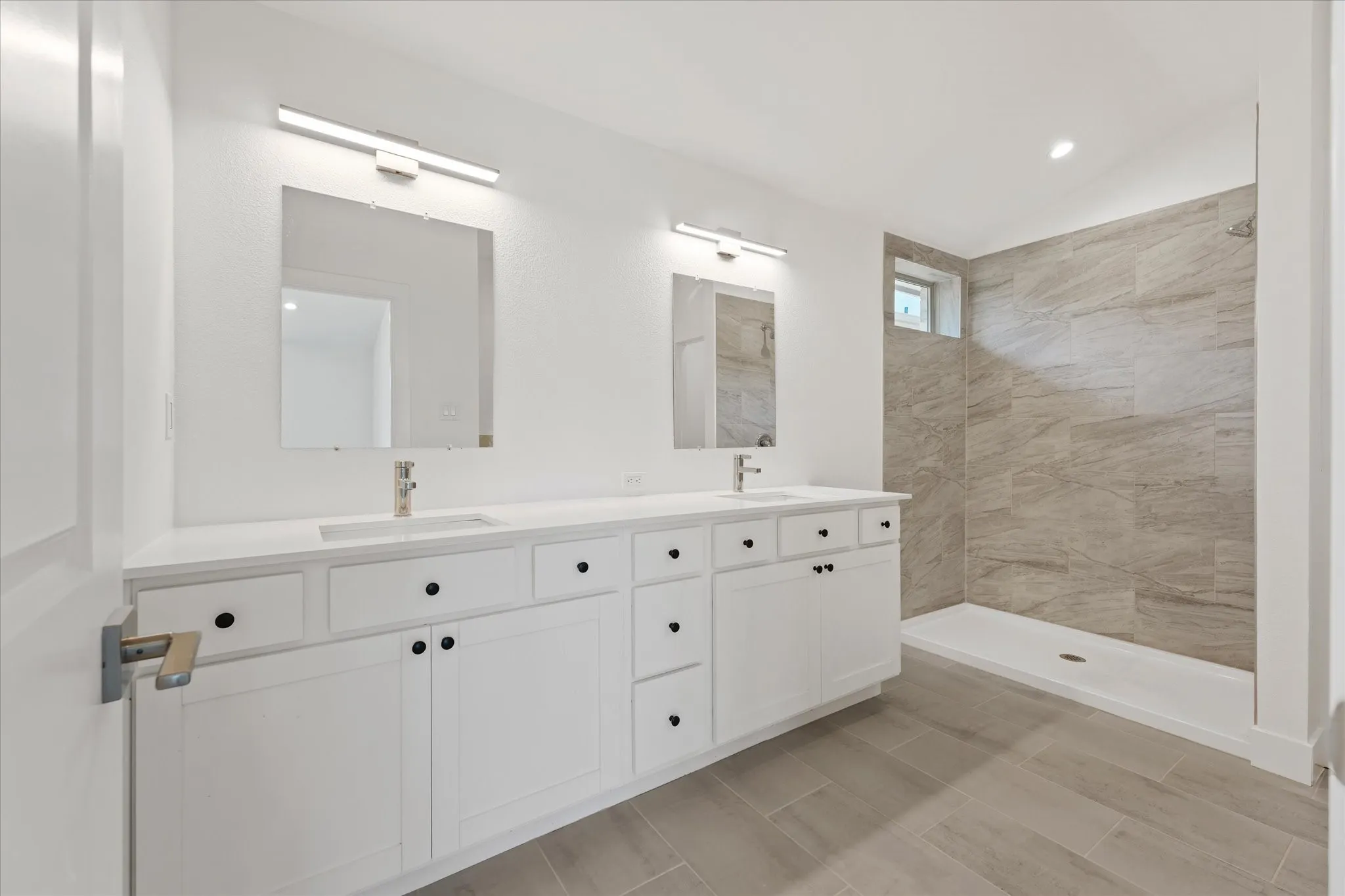 Dual vanity with white cabinetry and black hardware, quartz countertop, two rectangular undermount sinks, and a walk-in shower with tile surround and a transom window