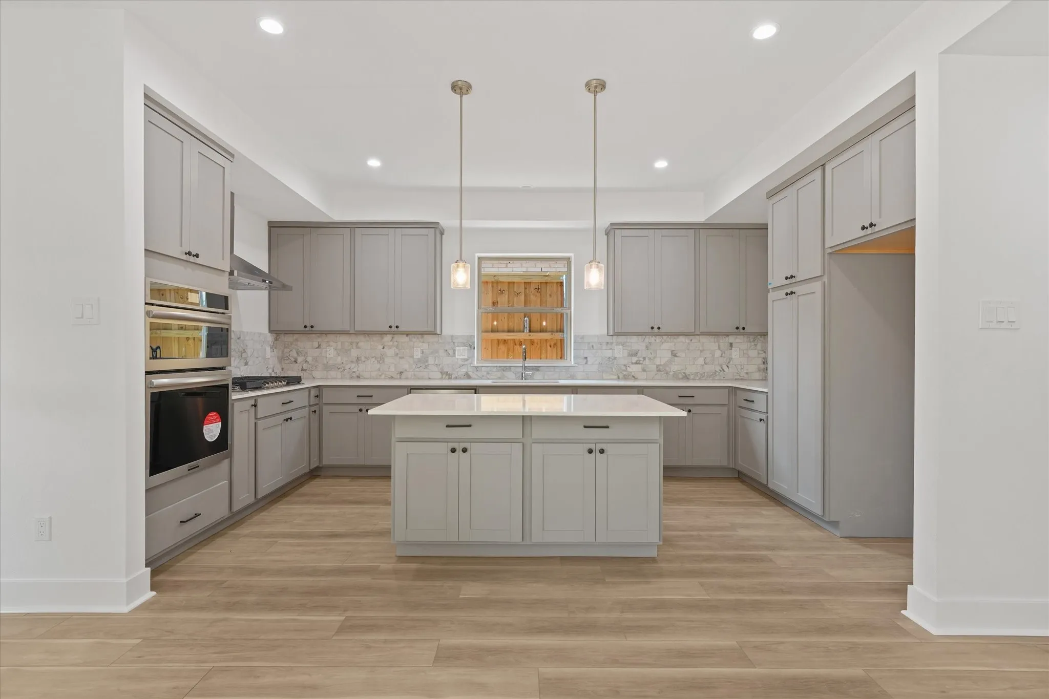 Modern kitchen featuring an island, light gray cabinetry, stainless steel appliances, wood-finish flooring, and pendant lighting