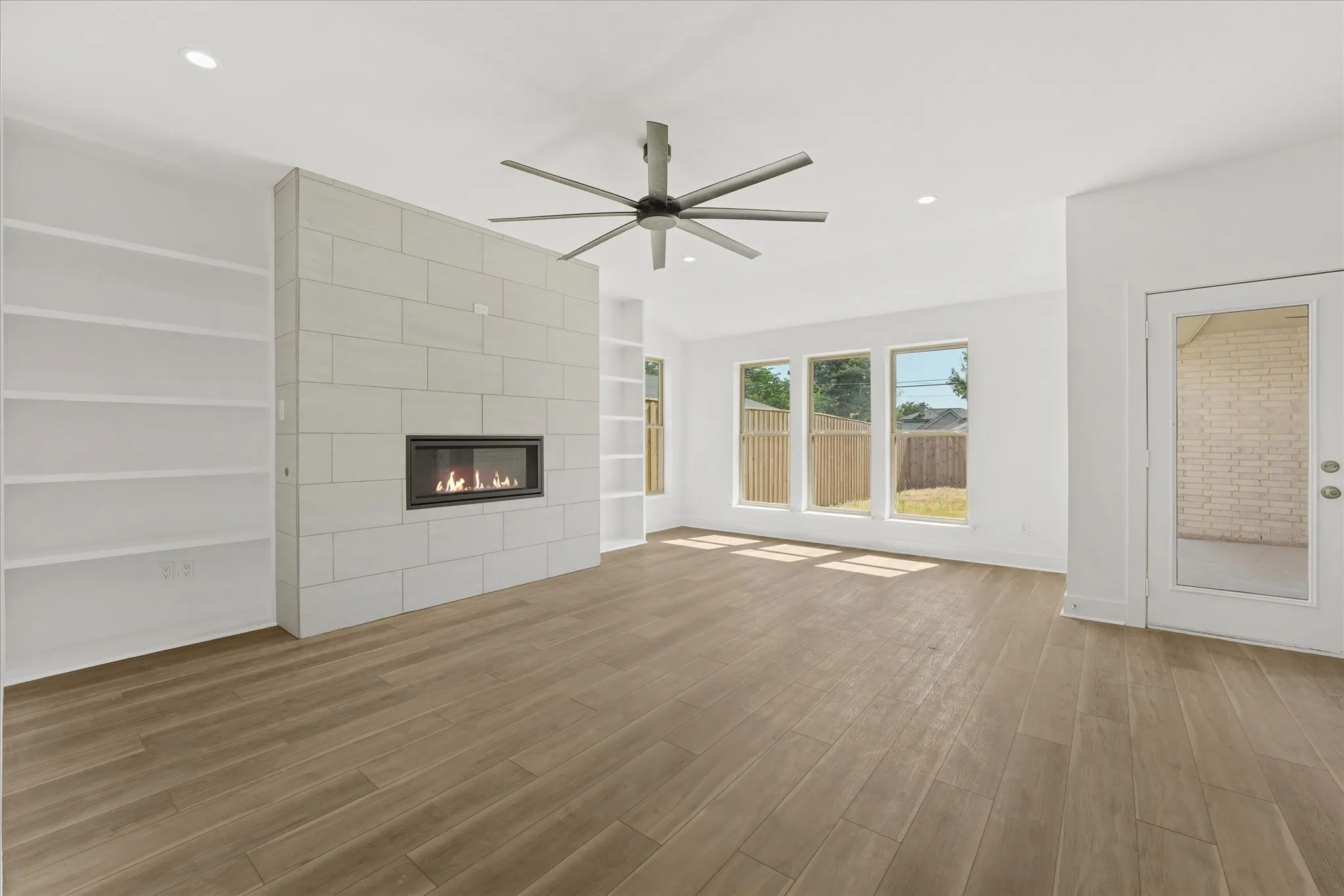 Wood-finish flooring, built-in shelving, linear fireplace with tile surround, ceiling fan, and multiple windows