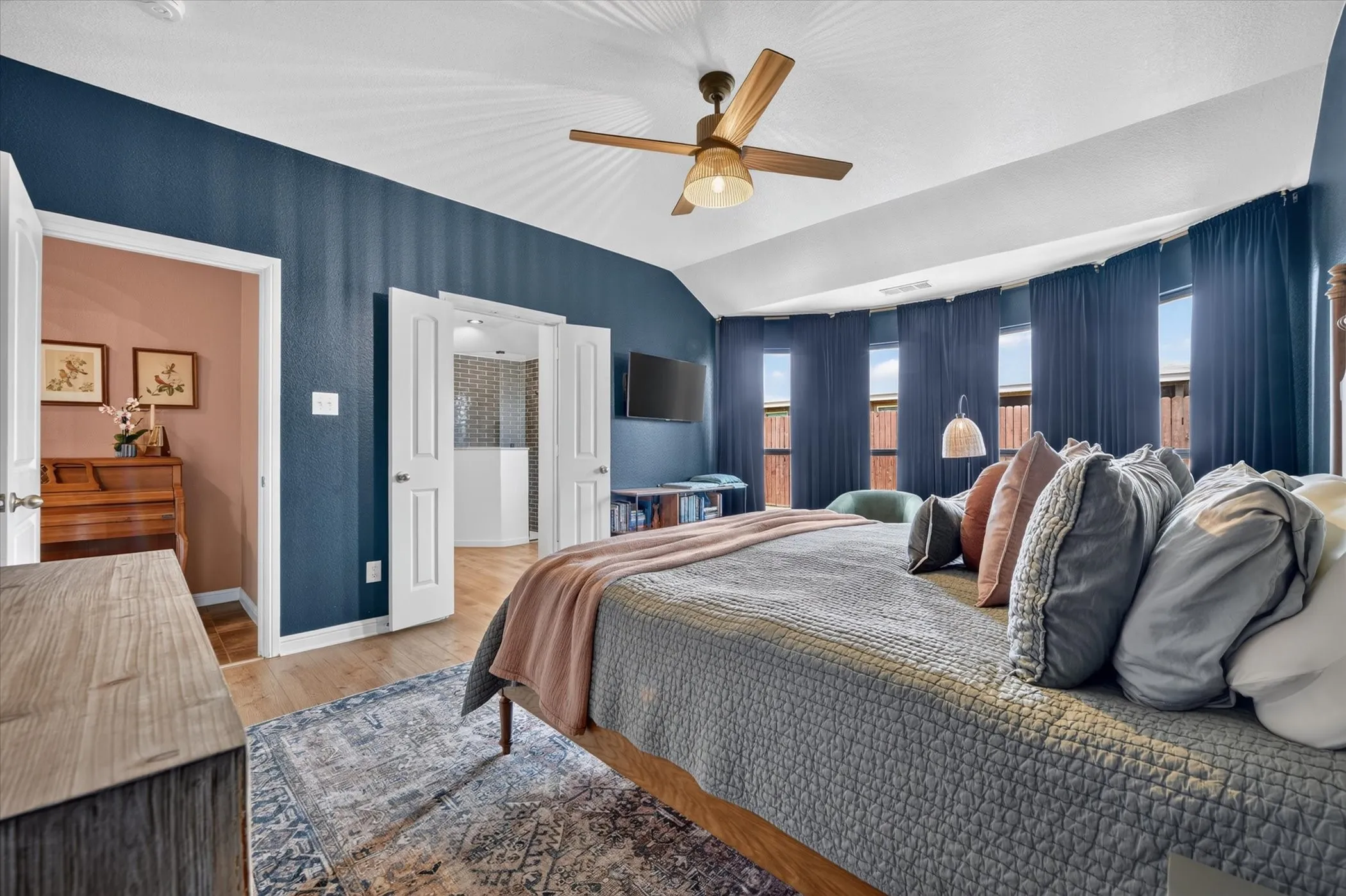 Vaulted ceiling with wood-finish ceiling fan, light wood-finish flooring, and multiple windows with full-length drapery