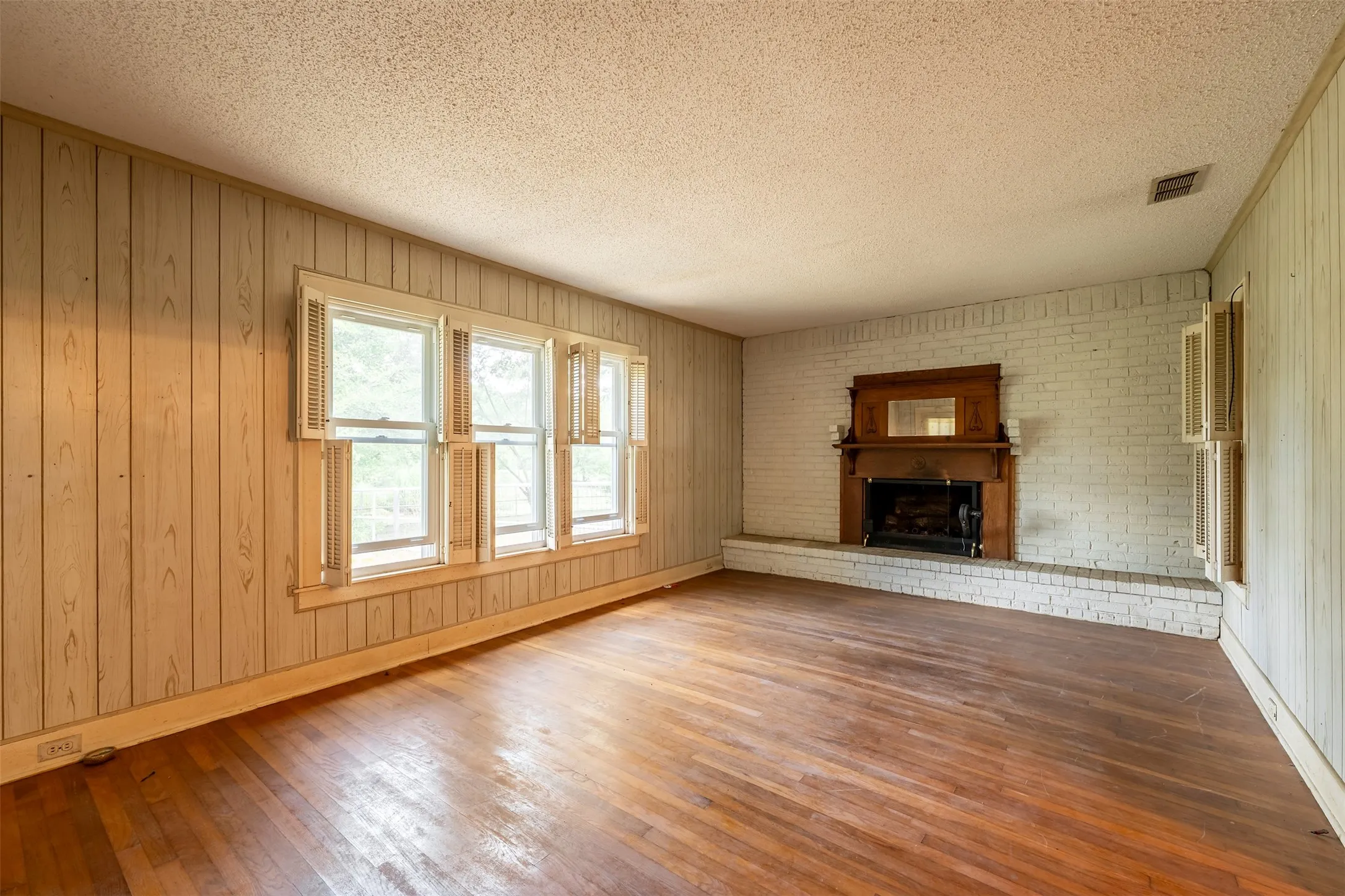 Wood-finish flooring, white painted brick fireplace with a wood mantel, multiple windows with shutters, and wood-paneled walls