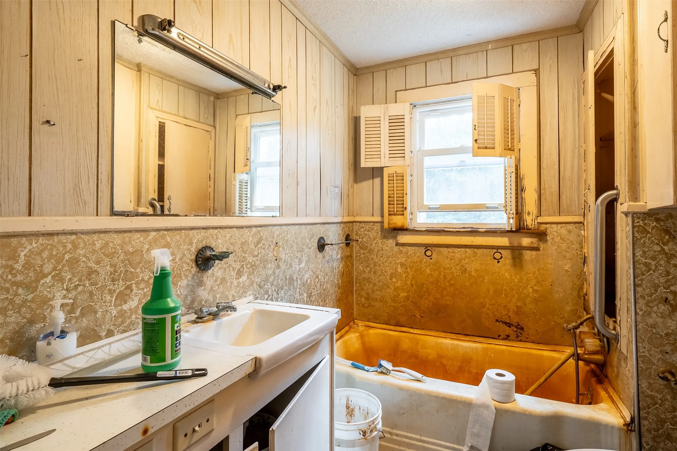 Wall-mounted vanity mirror, single window with interior shutters, wall paneling, built-in bathtub, and a vanity cabinet with a sink