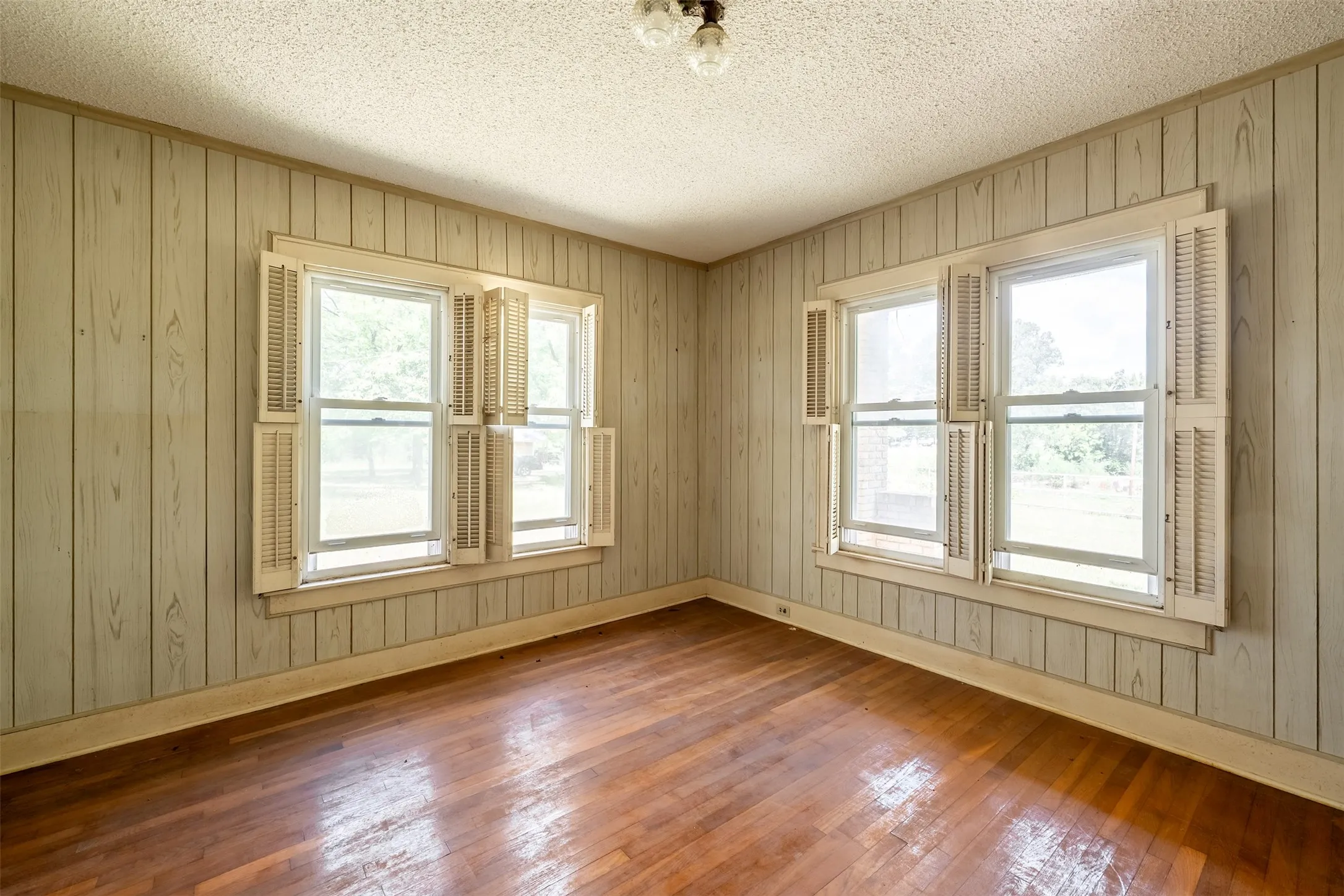 Wood-finish flooring with a sheen, vertical wood paneling, multiple windows with interior shutters, and a textured ceiling