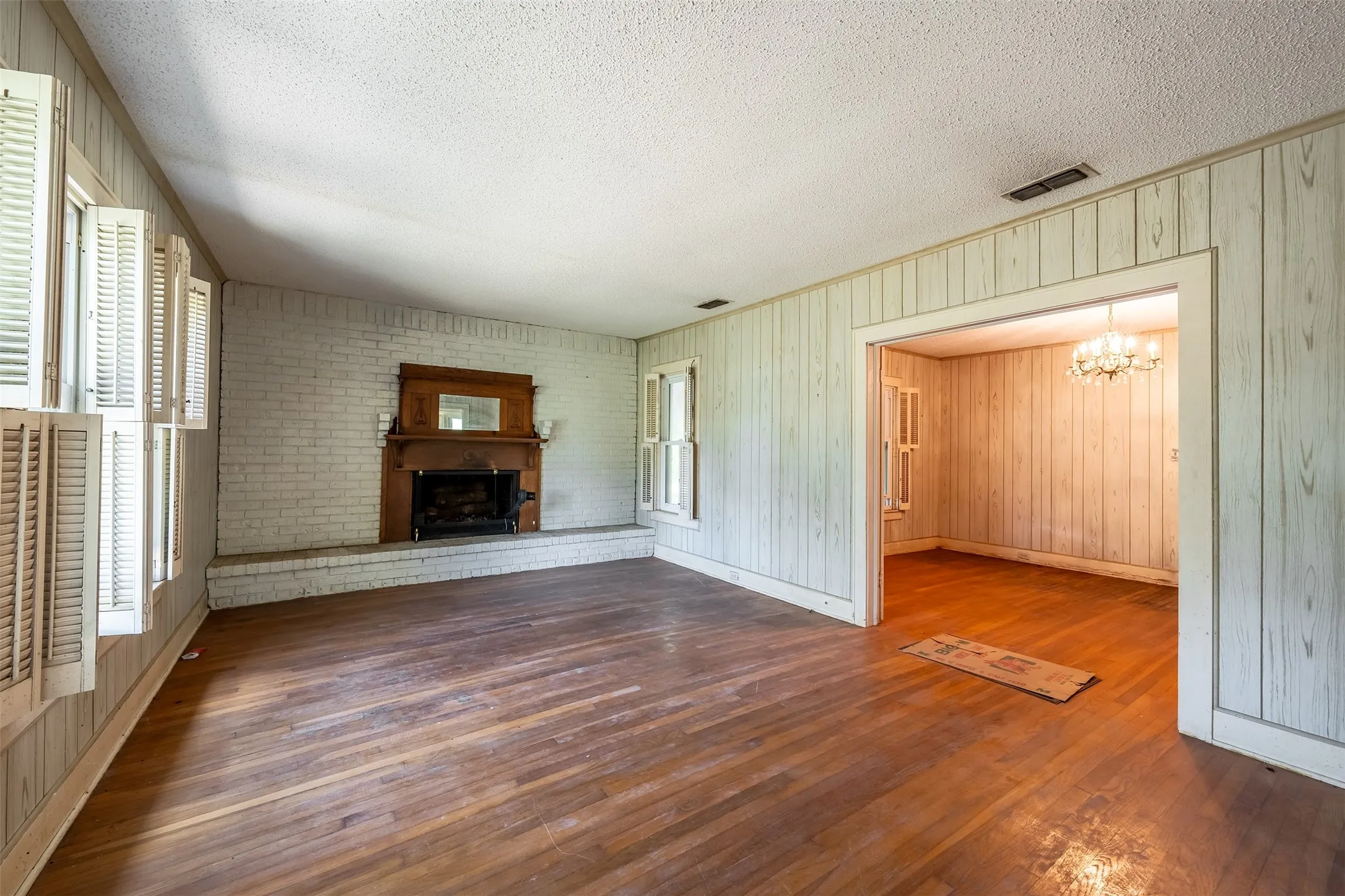 Hardwood flooring, brick fireplace with wood mantel, paneling, open doorway, and a chandelier in the adjacent space