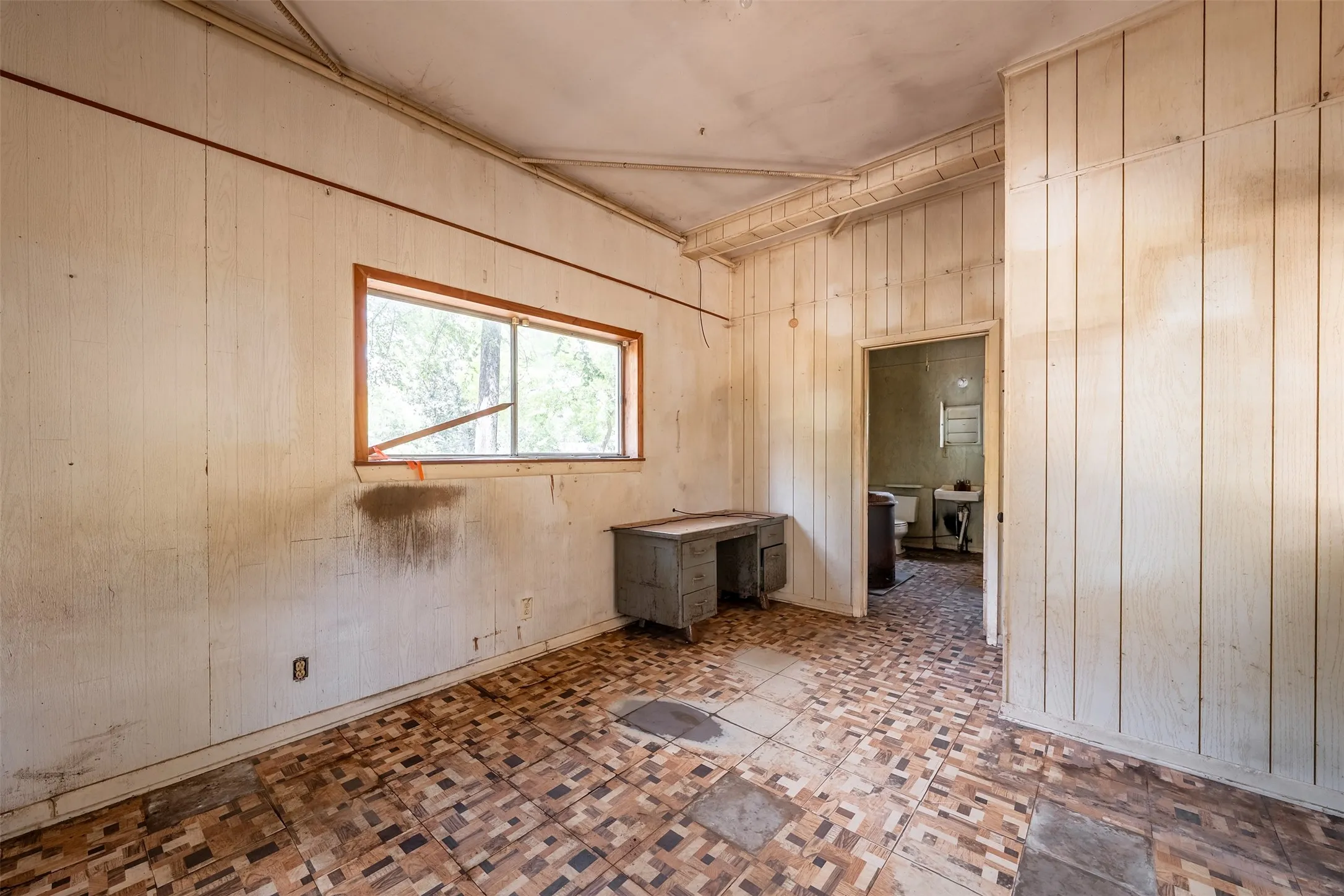 Spacious interior room featuring original wood-finish flooring in a classic brick pattern, wood-paneled walls, and a large fixed window with wood framing that provides natural light