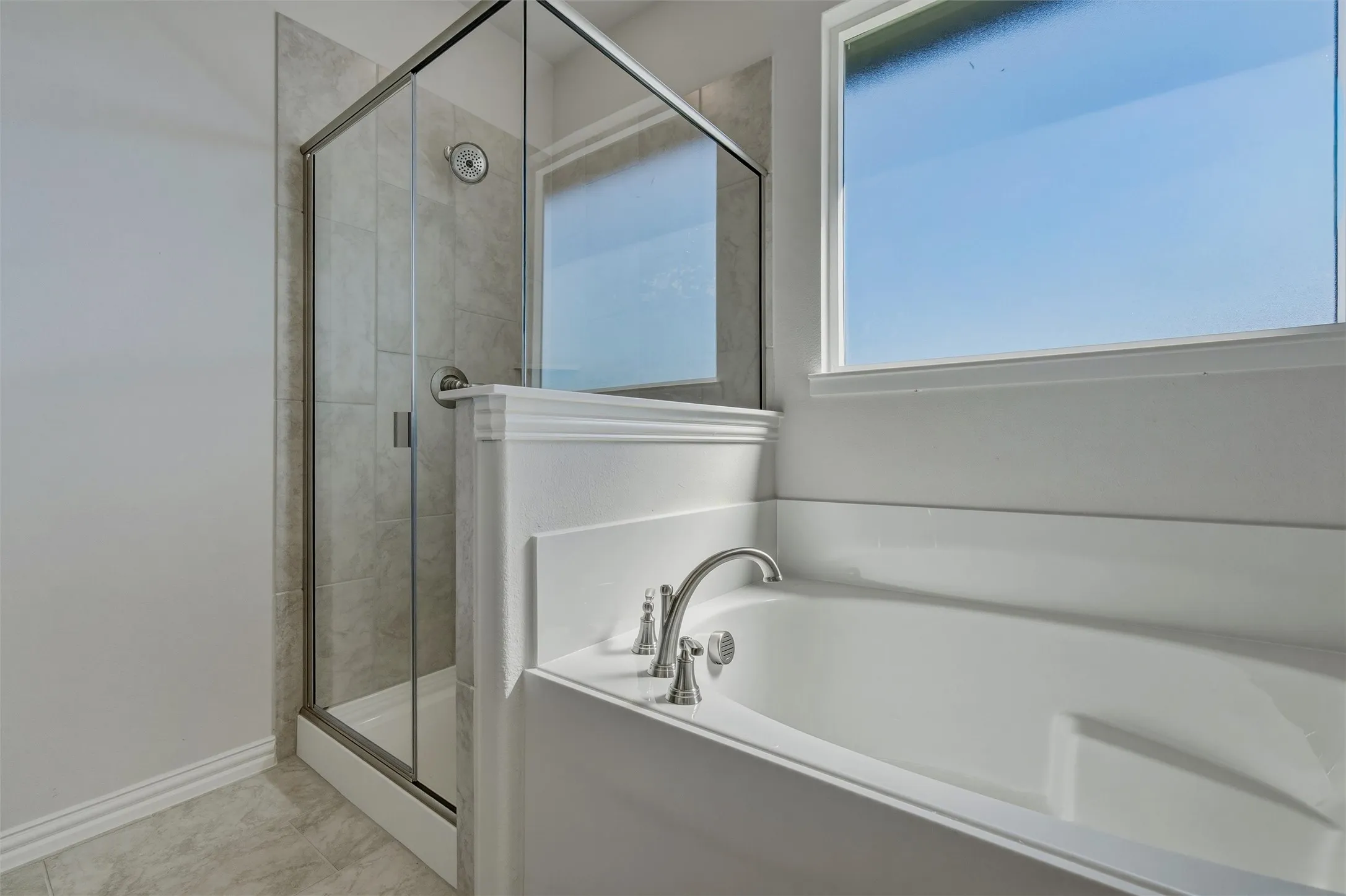 Soaking tub with brushed nickel fixtures, tiled shower with glass enclosure, white window framing, and light-toned floor tiling