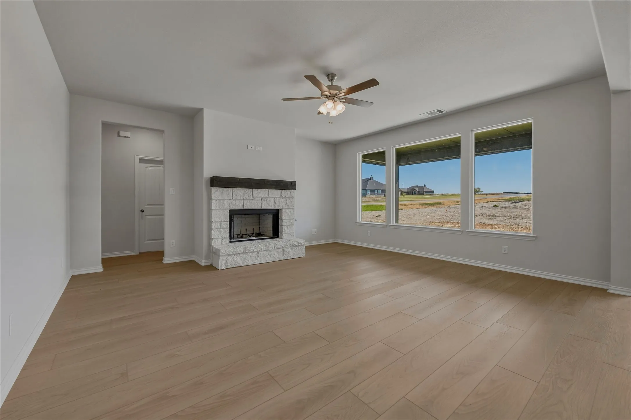 Spacious living area featuring wood-finish flooring, a stone fireplace with a dark wood mantel, multiple large windows, and a ceiling fan