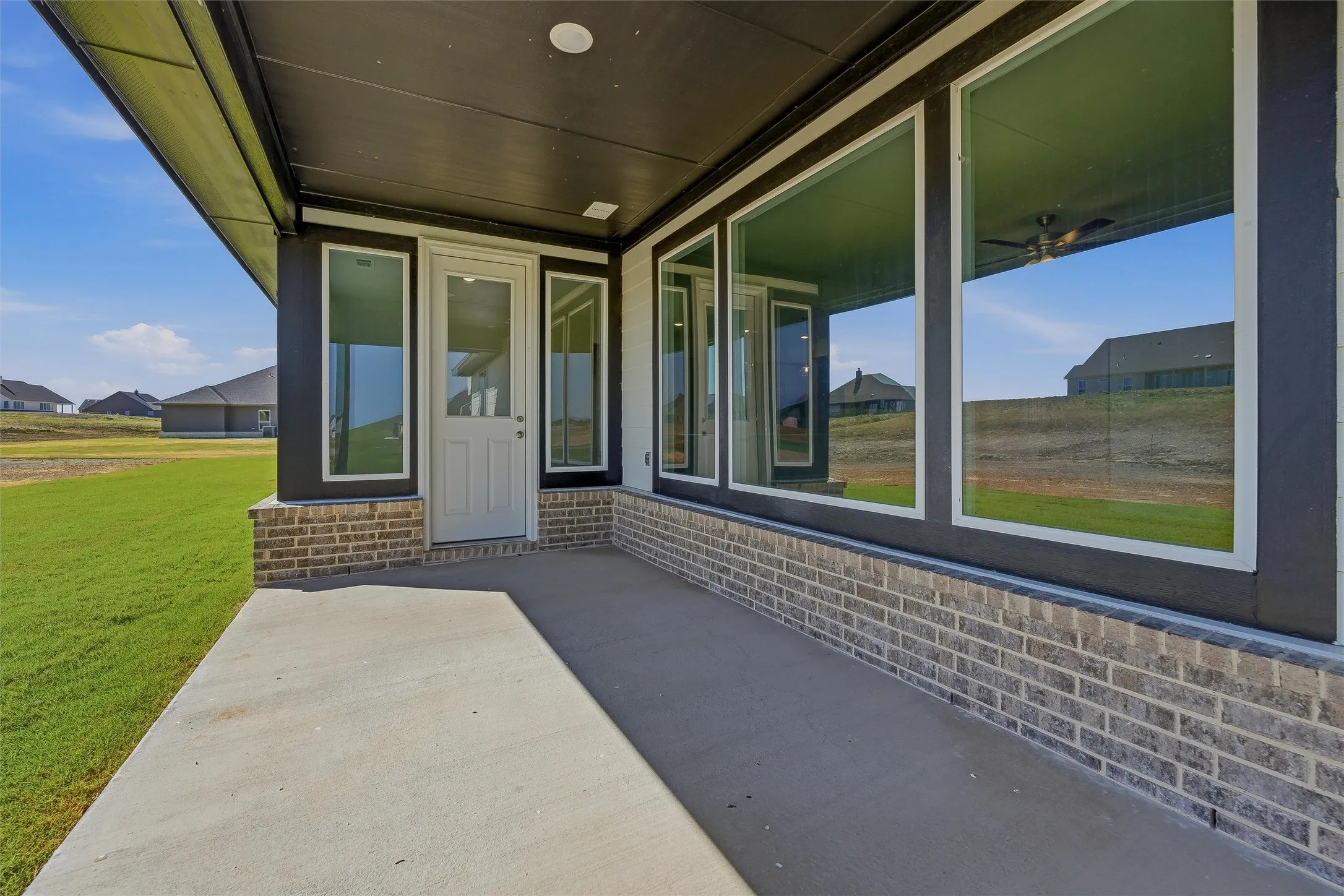 Covered porch with expansive windows framing pastoral landscapes, brick and stone exterior detailing, concrete flooring, and a white entry door with sidelights—ideal for indoor-outdoor entertaining and natural light throughout