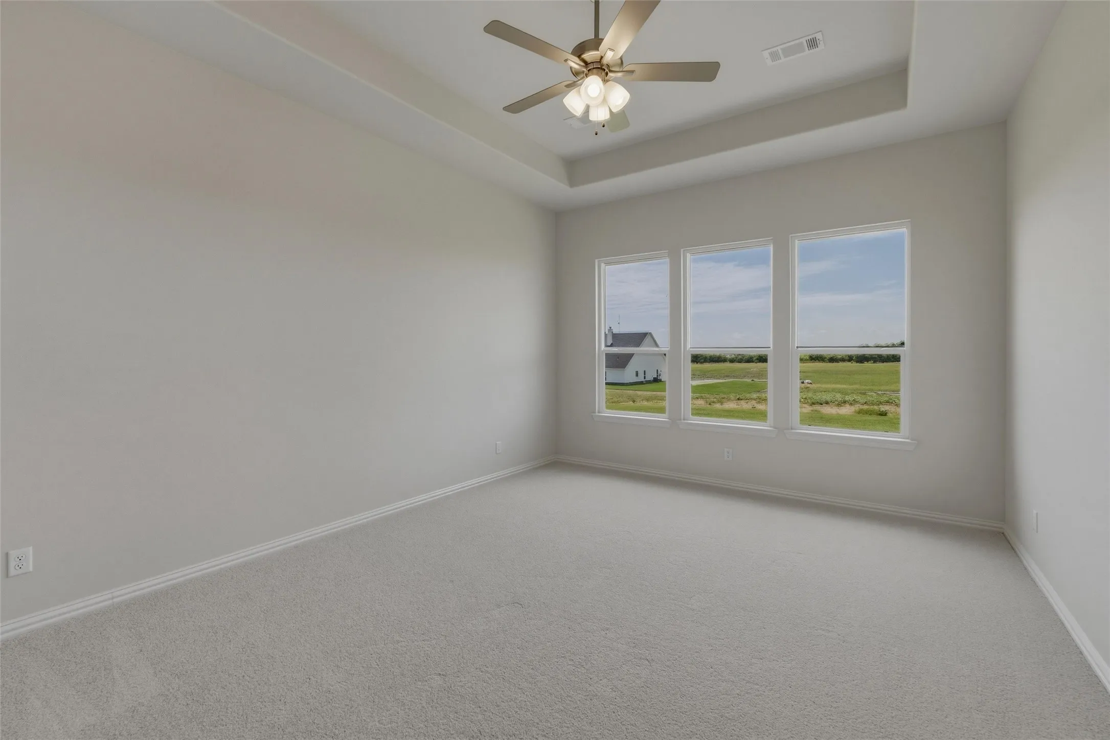 Spacious room featuring a tray ceiling, ceiling fan with integrated lighting, three large windows, and light gray carpeting