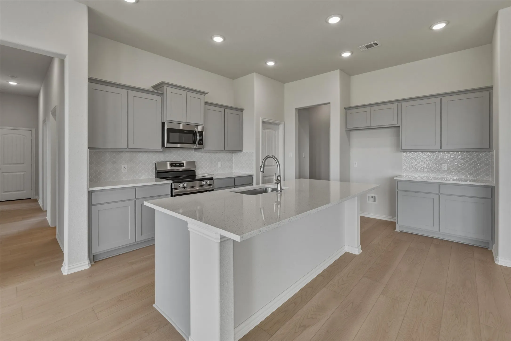 Kitchen featuring an expansive island with a sink, light grey cabinetry, stainless steel appliances, decorative backsplash, and wood-finish flooring
