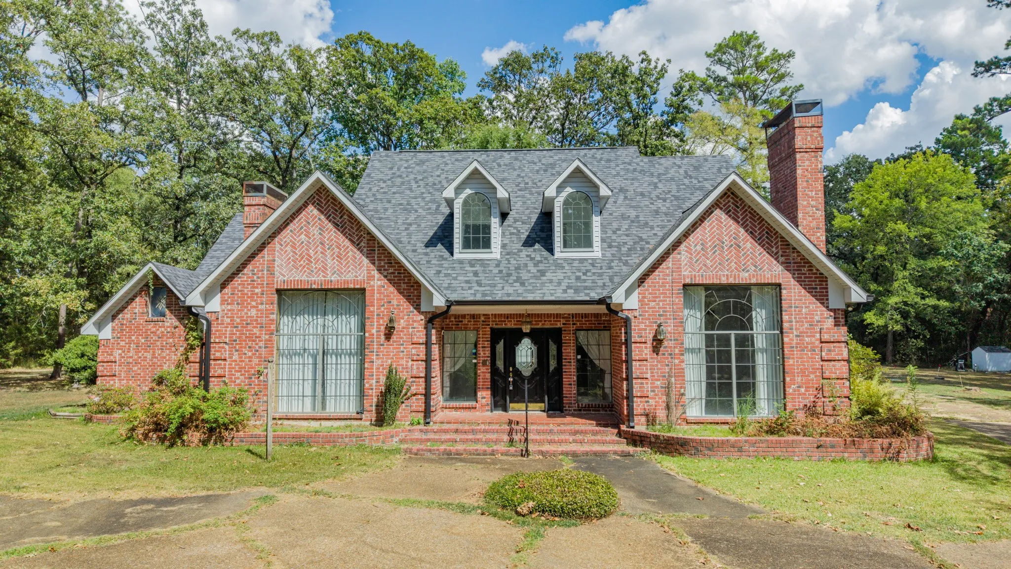 Brick exterior with a gray shingle roof, twin chimneys, a covered entryway, and arched dormer windows