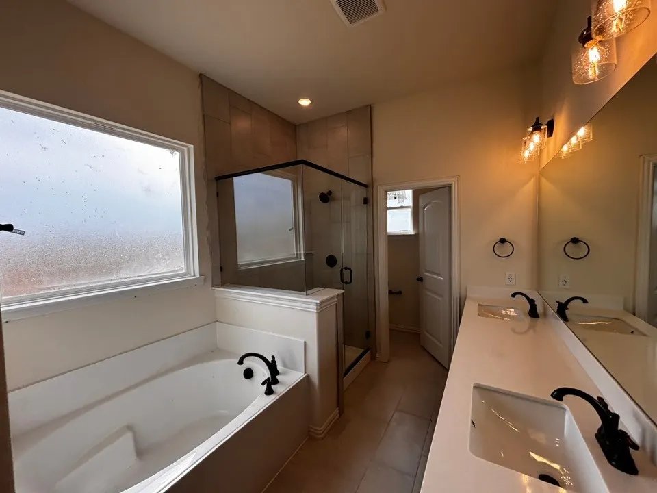 Dual vanity with integrated sinks, frameless glass shower, built-in bathtub, tile flooring, and decorative vanity lighting