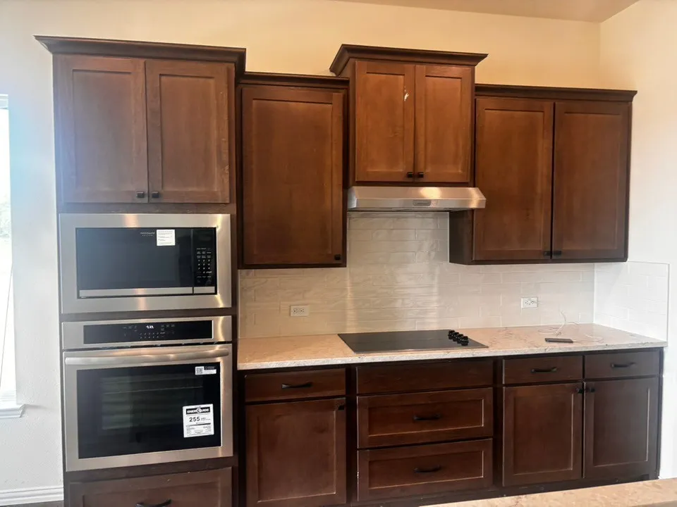 Kitchen featuring rich wood cabinetry, stainless steel built-in microwave and oven, a sleek cooktop, light-toned countertops, and a subway tile backsplash