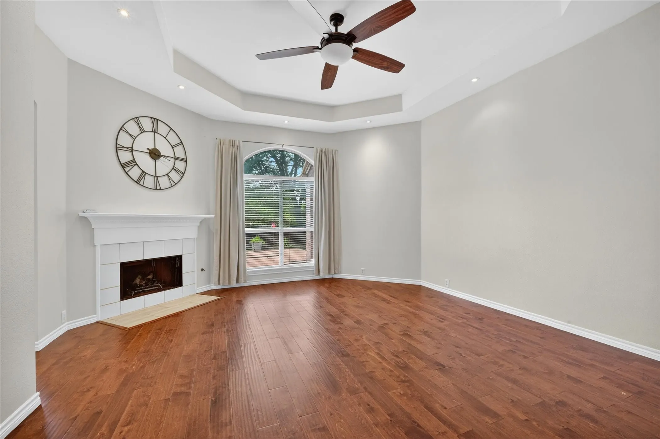 Wood-finish flooring, fireplace with tile surround, arched window, tray ceiling, and recessed lighting