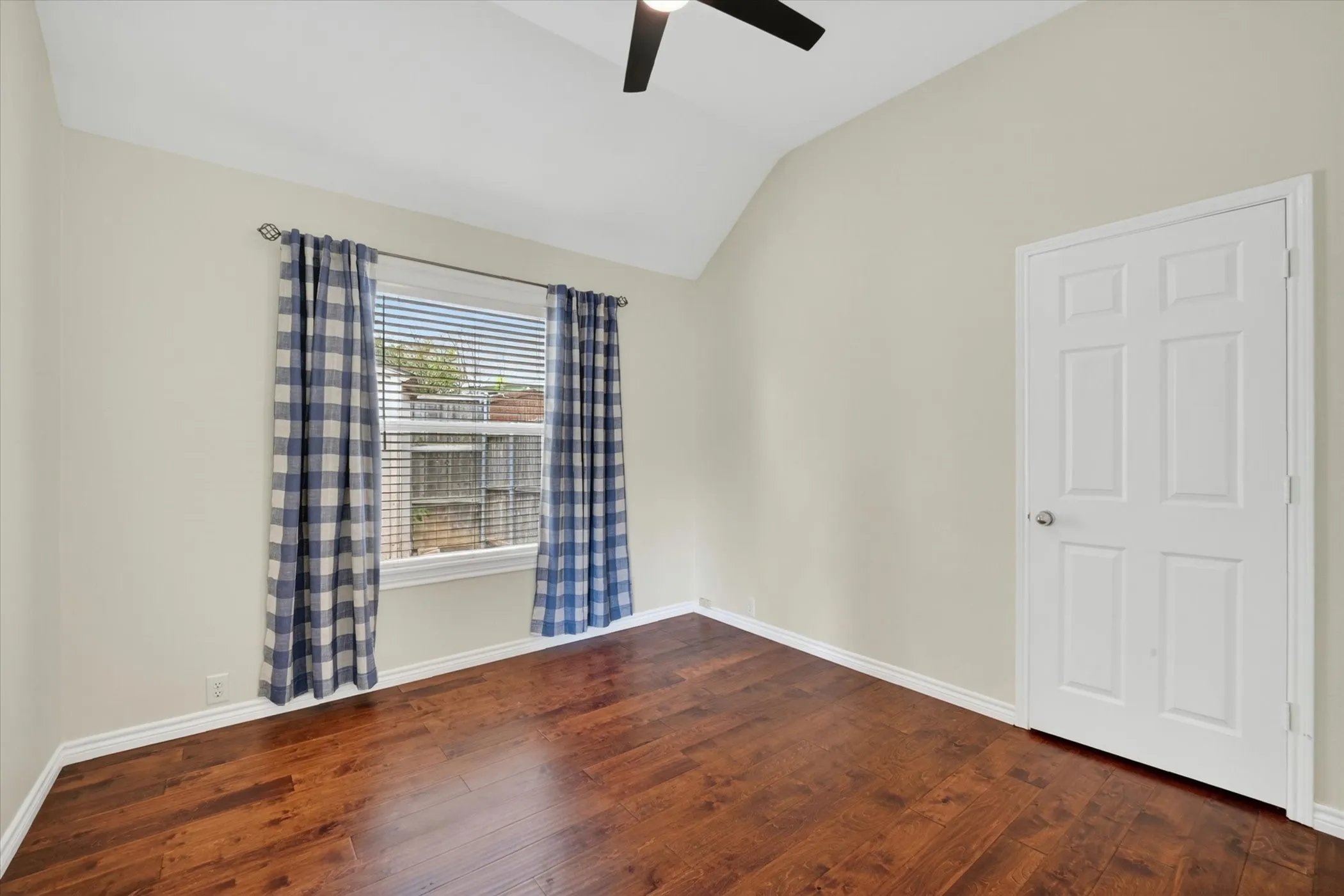 Bright bedroom showcasing hardwood flooring, vaulted ceiling with ceiling fan, and large window with blue plaid curtains