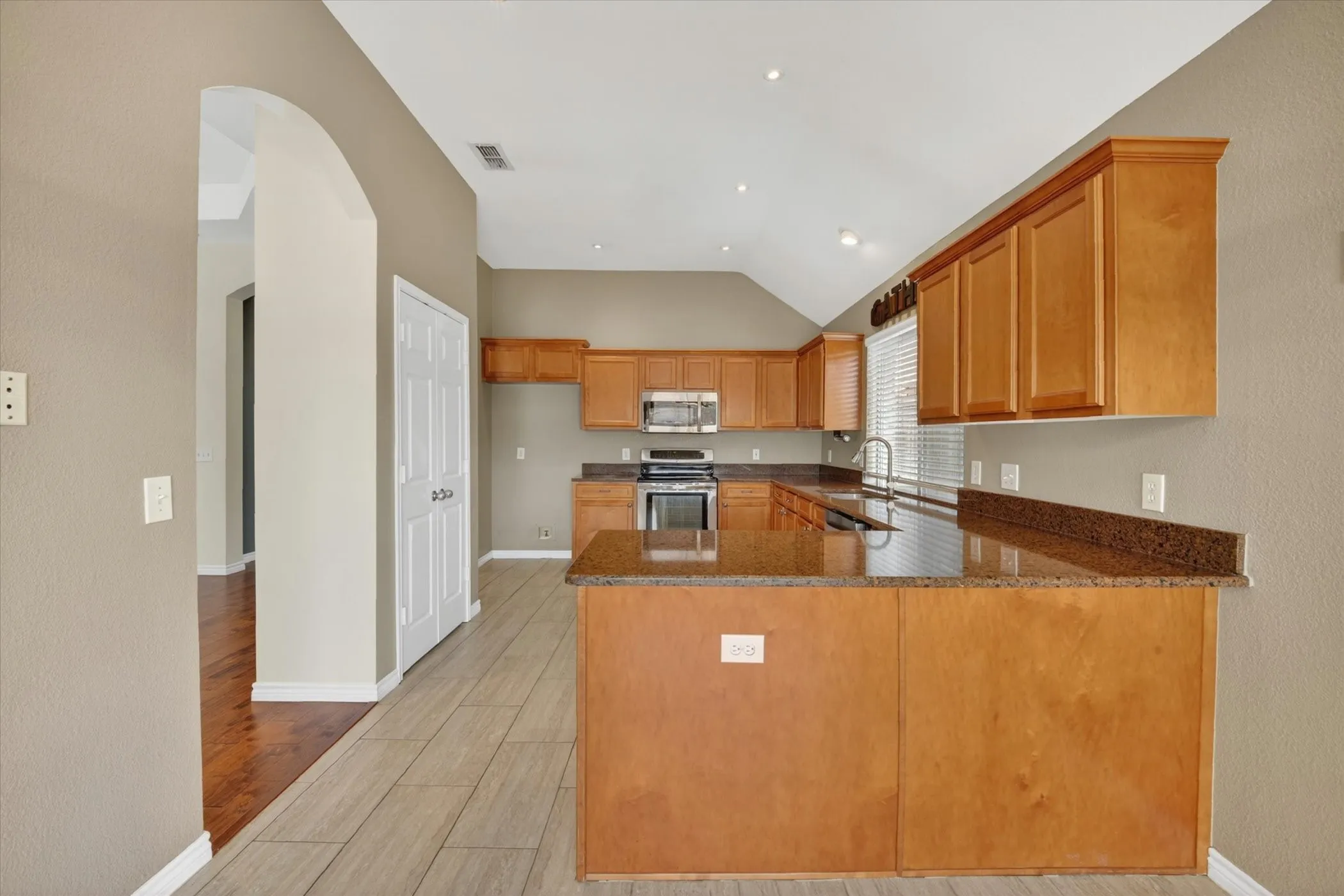 Spacious kitchen featuring rich wood cabinetry, stainless steel appliances, granite countertops with waterfall island edge, subway tile backsplash, and wood-finish flooring throughout the open layout with vaulted ceiling and recessed lighting