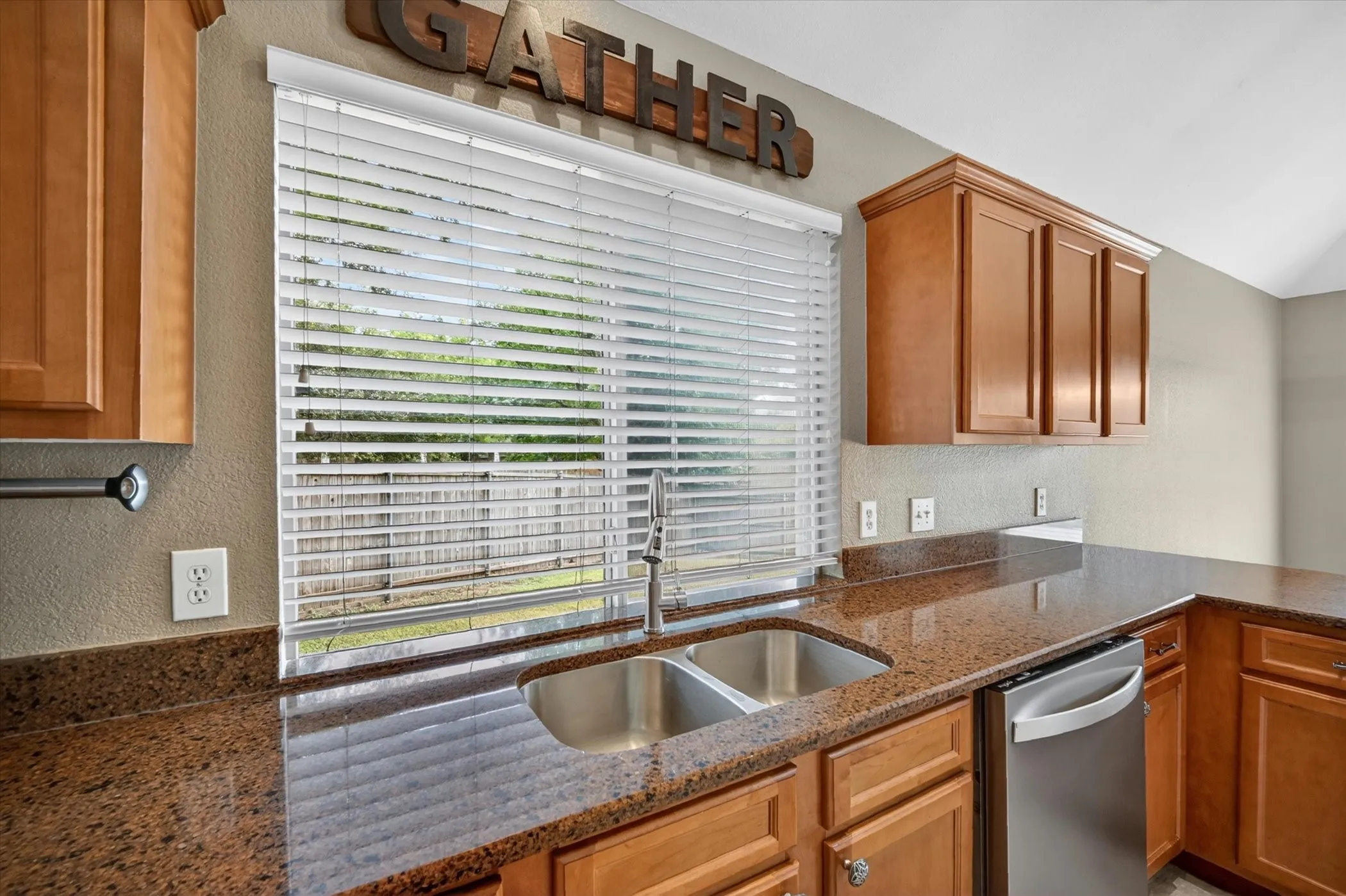 Spacious kitchen featuring warm wood cabinetry, granite countertops, stainless steel appliances, a double-basin sink, and expansive sliding glass doors with horizontal blinds overlooking outdoor space