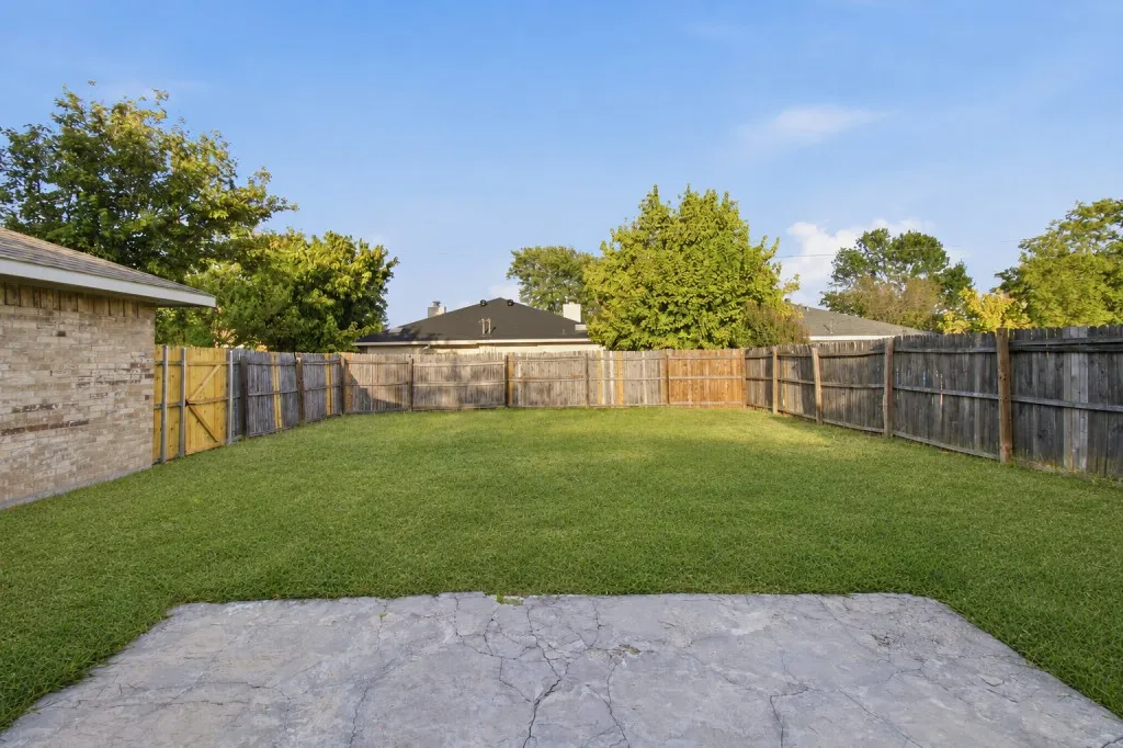Expansive backyard oasis featuring a level lawn with mature landscaping, wood-finish fencing with yellow-trimmed gate, and a concrete patio foundation