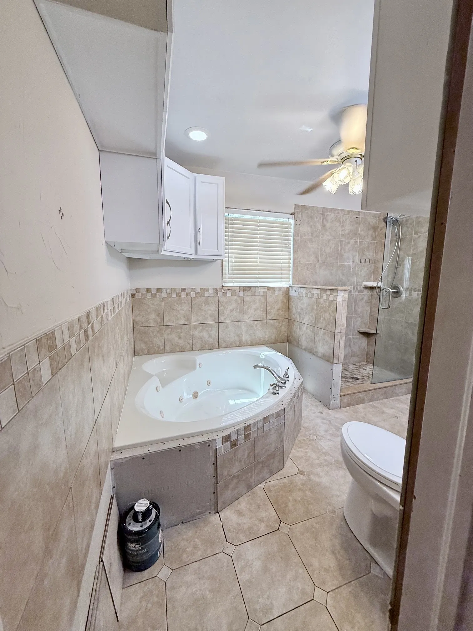 Corner jetted soaking tub, glass-enclosed walk-in shower, tile flooring, and integrated ceiling fan