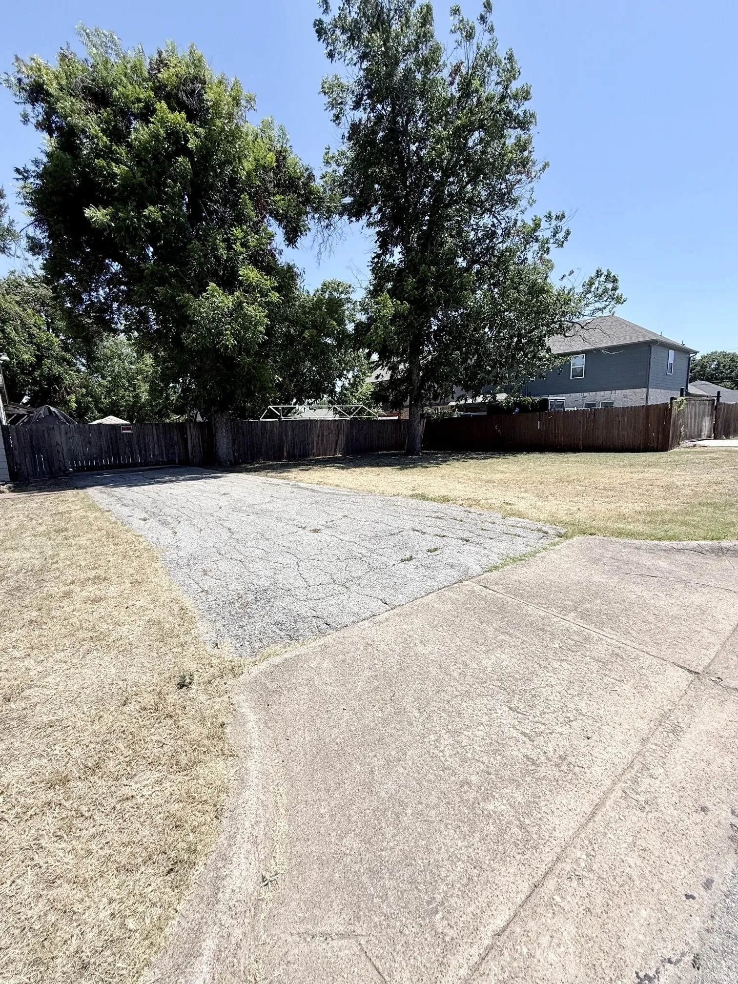 Expansive lot featuring mature trees, a wooden fence, and concrete paving