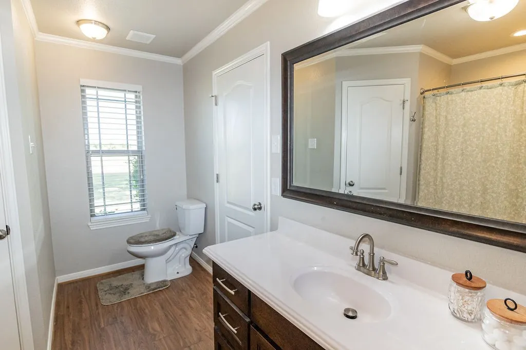 Wood-finish flooring, single vanity with a white countertop and integrated sink, framed mirror, and a window with blinds