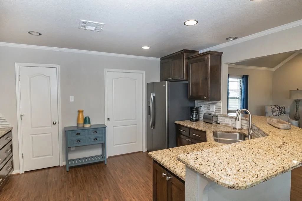 Contemporary kitchen featuring granite-style countertops, a large center island with seating space, stainless steel appliances including a refrigerator and gas cooktop, dark wood cabinetry, subway tile backsplash, recessed ceiling lighting, and wood-finish flooring throughout the open layout