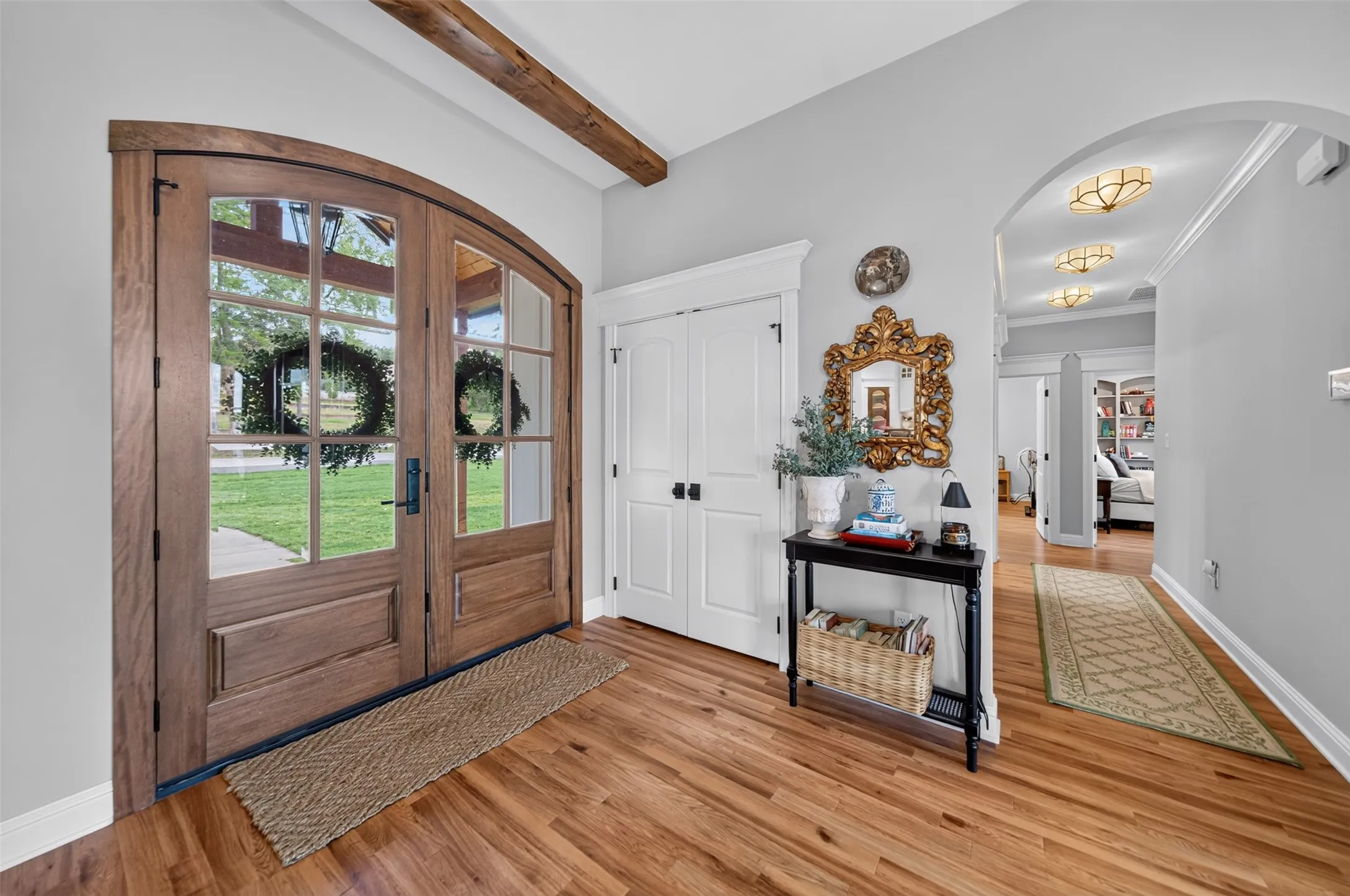 Arched double entry doors with divided lites, wood-finish flooring, exposed wood ceiling beam, and an arched hallway opening with crown molding