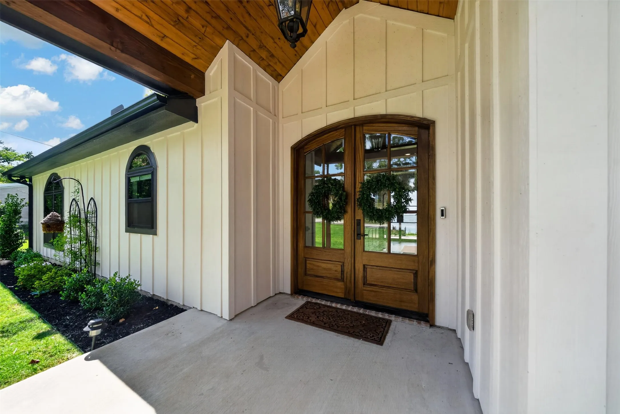 Arched wood-finish entry doors, board and batten siding, dark-framed arched windows, wood-paneled porch ceiling, and a concrete pathway
