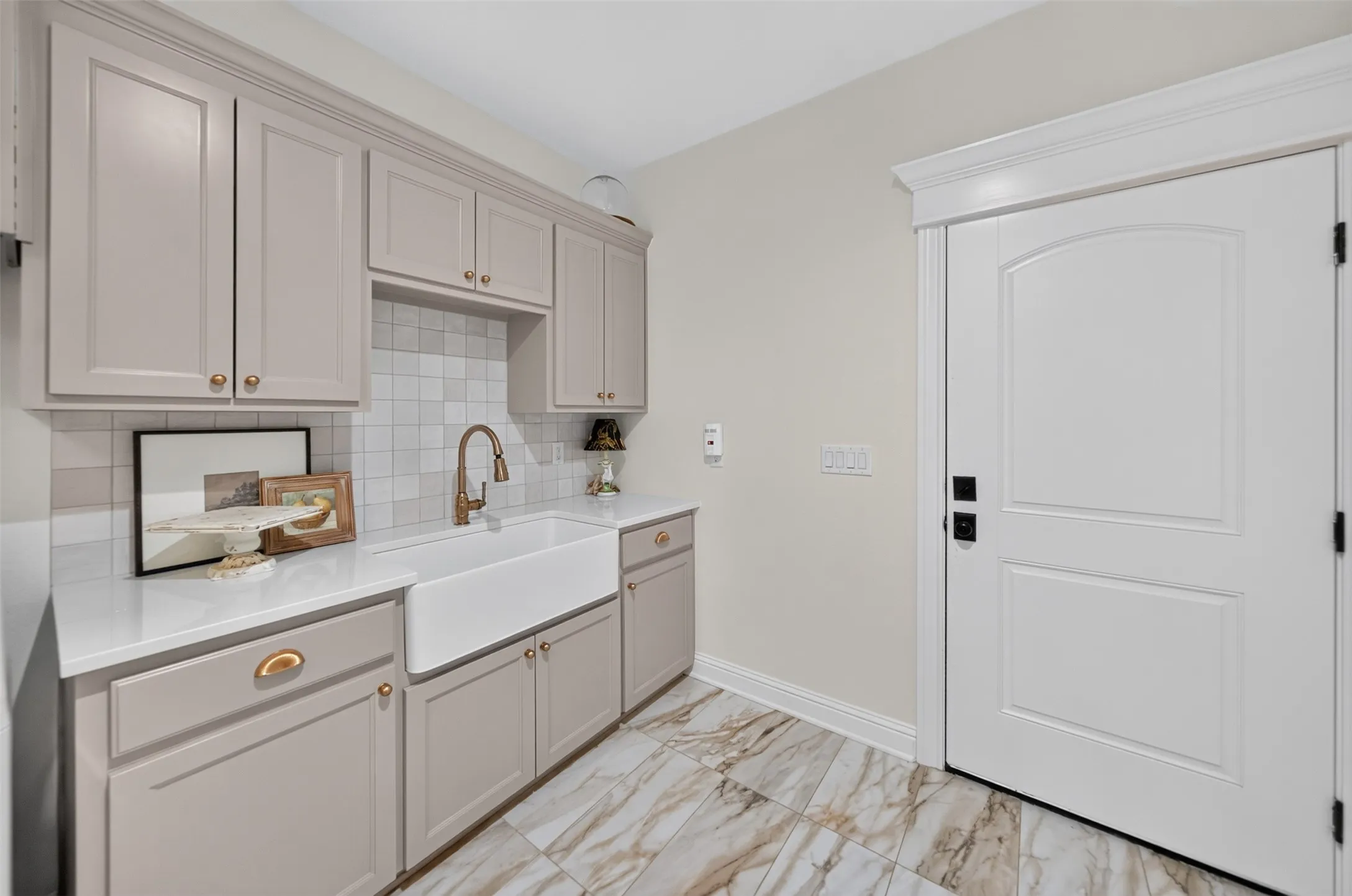Light-toned cabinetry with gold-toned hardware, solid surface countertops, white farmhouse sink with a gooseneck faucet, light-toned backsplash, and patterned tile flooring