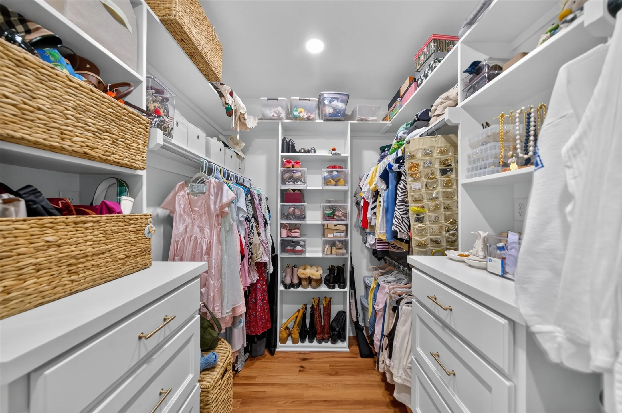 Custom closet system featuring white built-in cabinetry, warm wood-finish flooring, recessed lighting, and extensive shelving