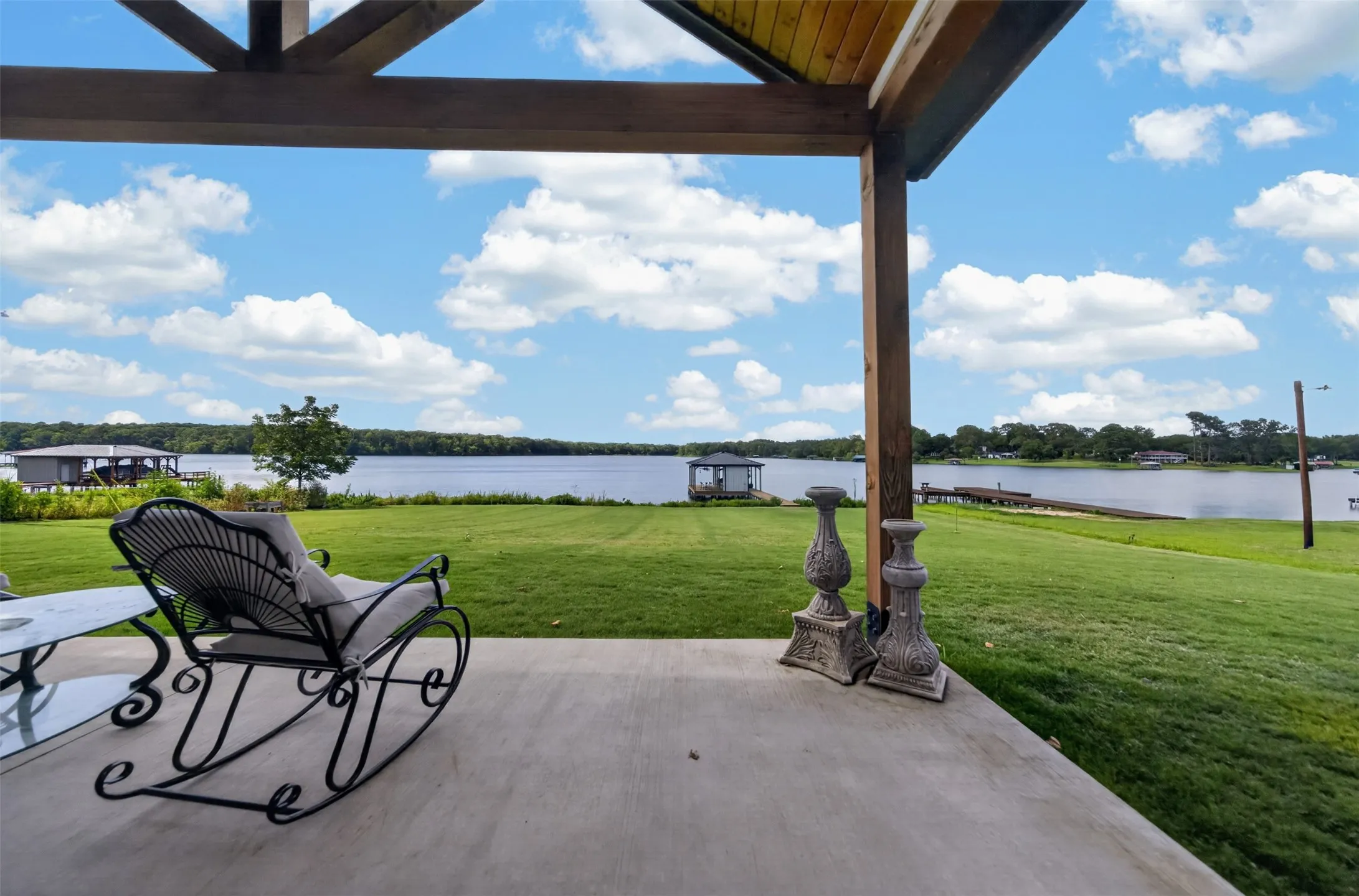 Expansive concrete patio with a wood-beamed overhead structure, overlooking a substantial body of water with a grassy shoreline