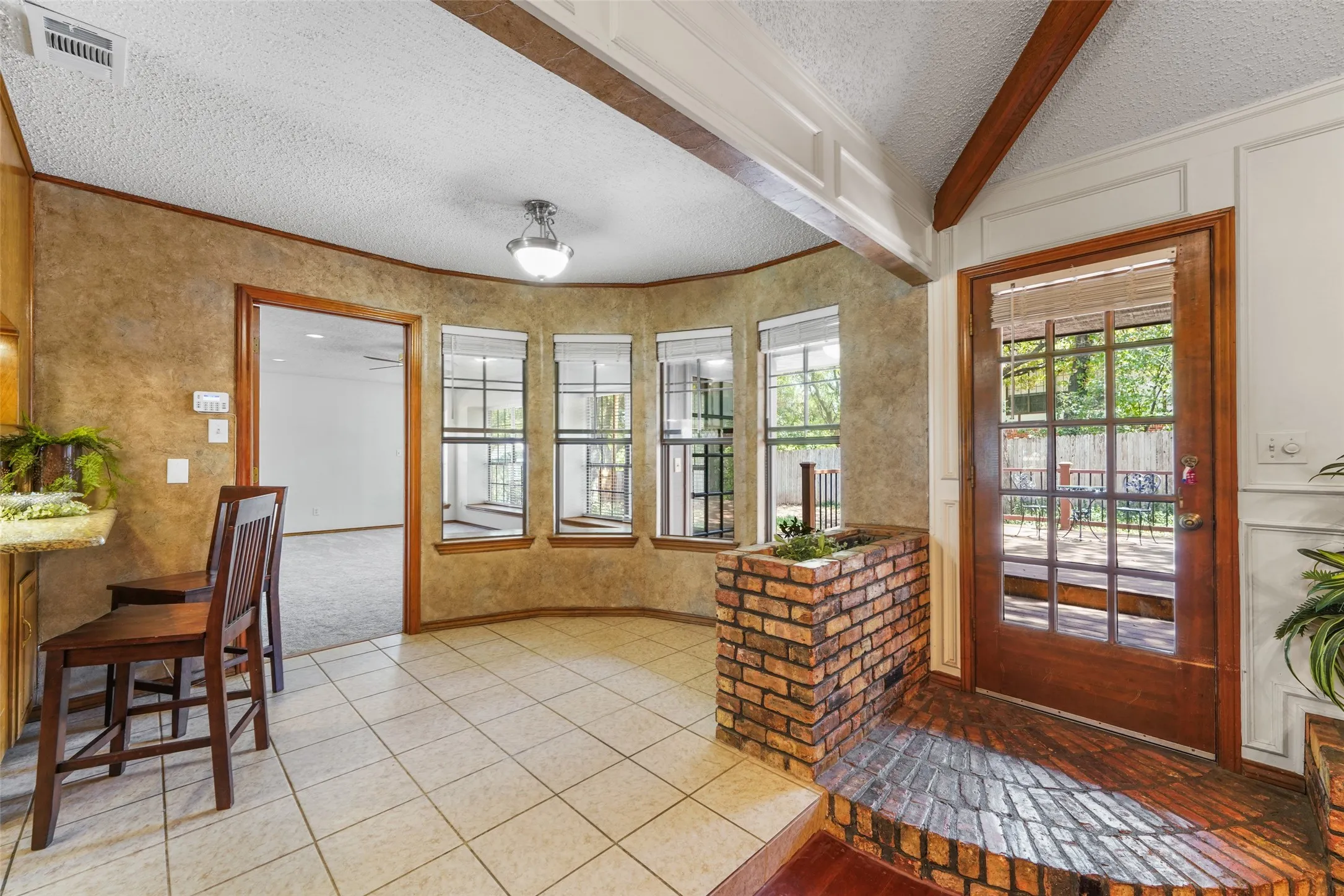 Bright, sun-filled kitchen and breakfast area featuring curved bay windows with wood frames, exposed beam ceiling detail, tile flooring, brick accent wall, and French doors opening to outdoor space