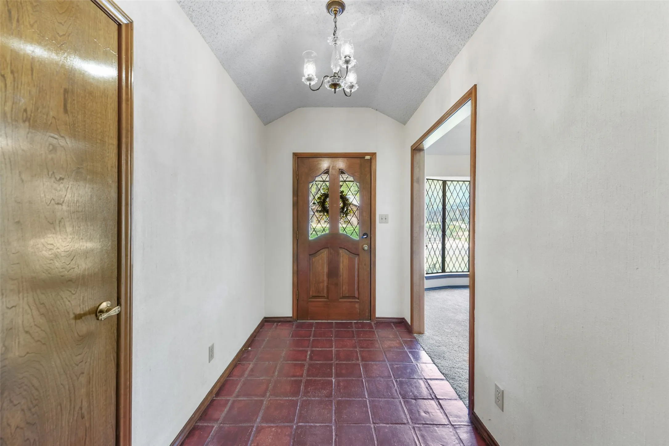 Vaulted ceiling entry with a three-light chandelier, wood entry door with decorative glass, and terracotta tile flooring