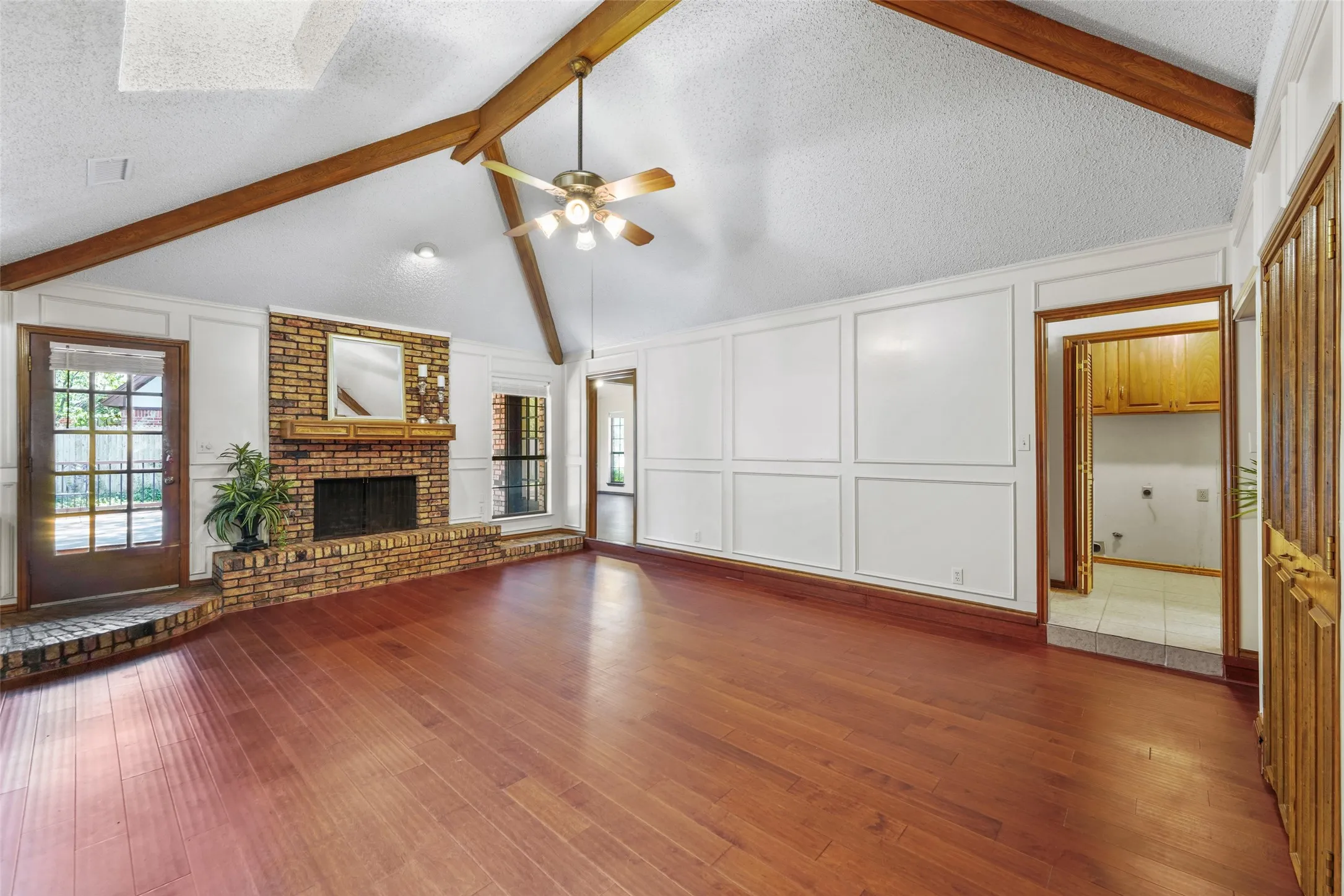 Vaulted ceiling with exposed wood beams, brick fireplace with raised hearth, wood-finish flooring, paneled wall detailing, and a ceiling fan