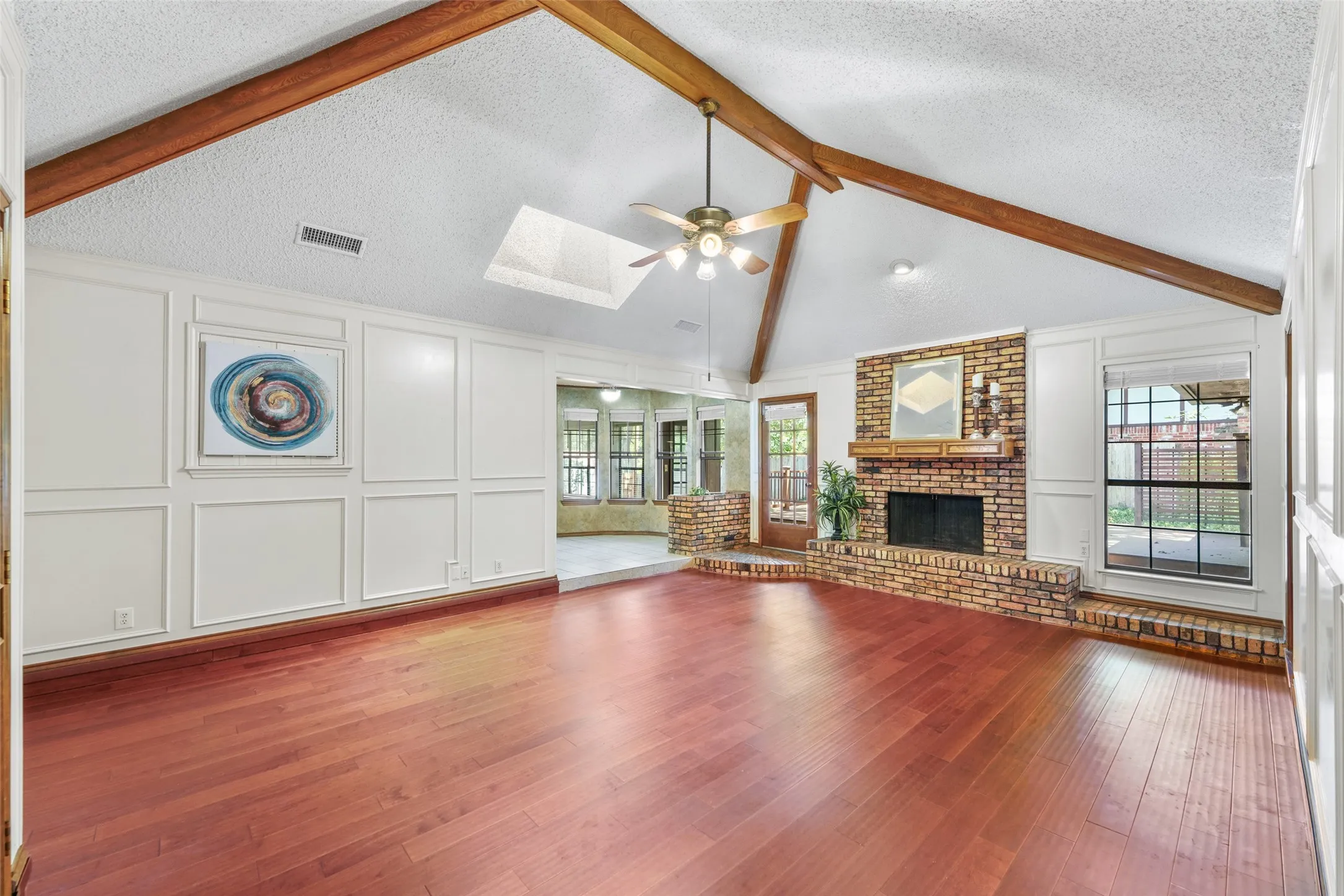 Vaulted ceiling with exposed wood beams, wood-finish flooring, brick fireplace with mantle, paneled accent wall, and a ceiling fan with integrated lighting