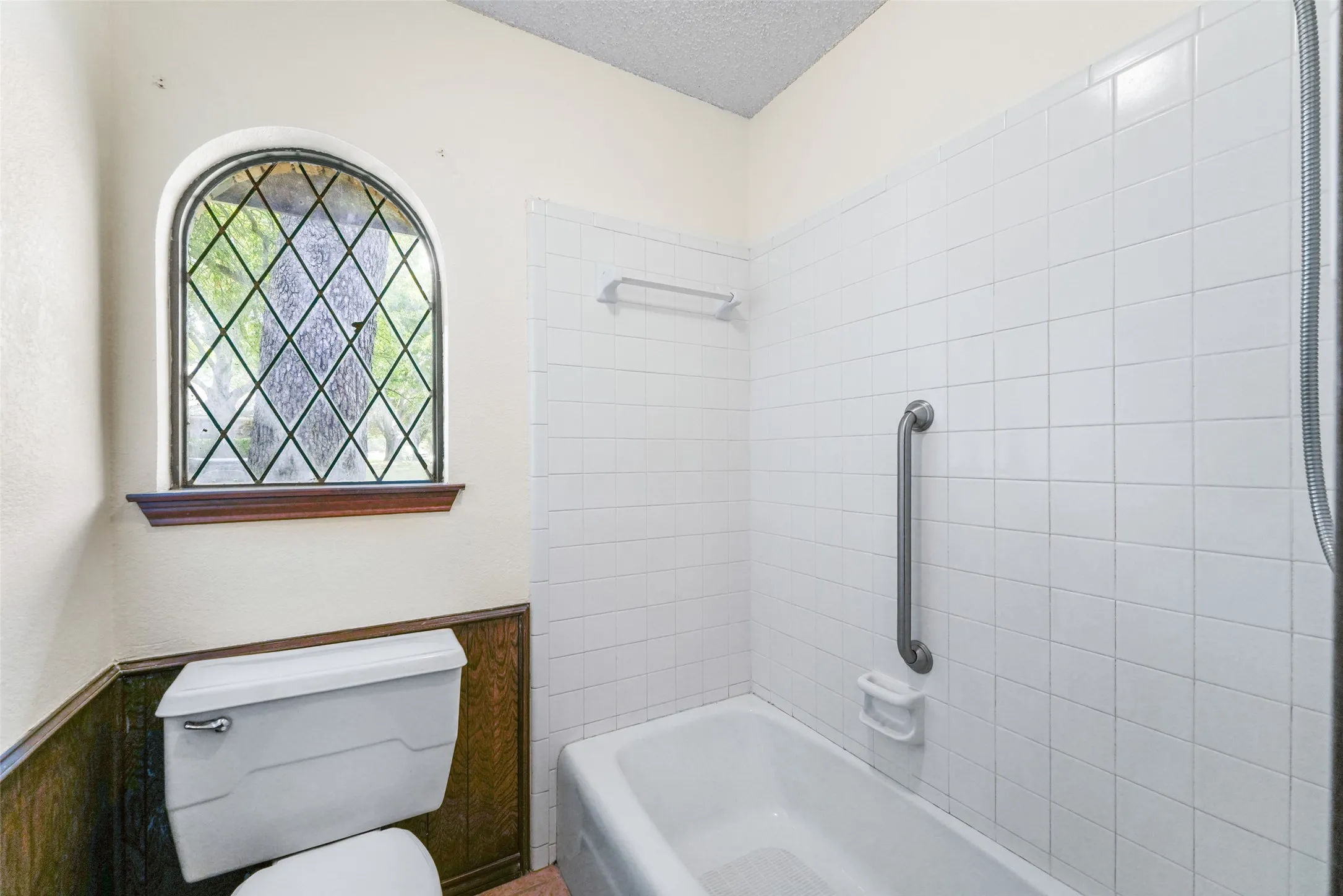 Bright bathroom featuring white subway tile walls, a classic white porcelain toilet with wood-finish cabinetry, and a white bathtub with chrome grab bar safety fixture