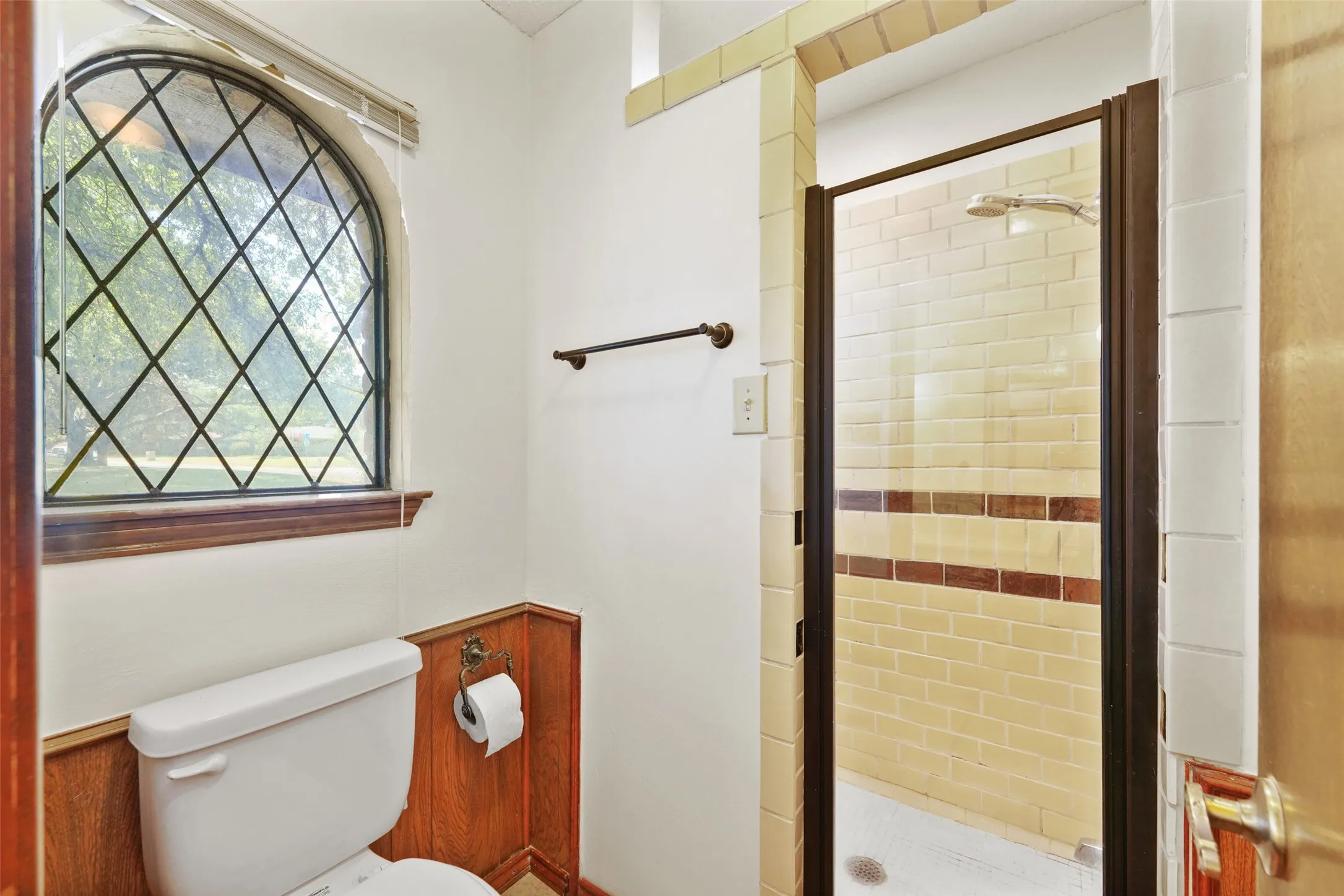 Arched window with diamond-patterned grid, wood-finish wainscoting, and a tiled shower with clear glass enclosure