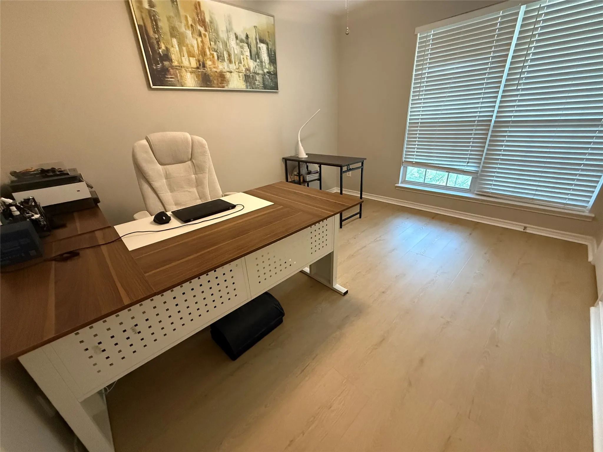 Professional office featuring a wood-finish desk with integrated storage, ergonomic upholstered chair, wood-finish flooring throughout, large window with horizontal blinds providing natural light, and neutral wall finishes with framed artwork